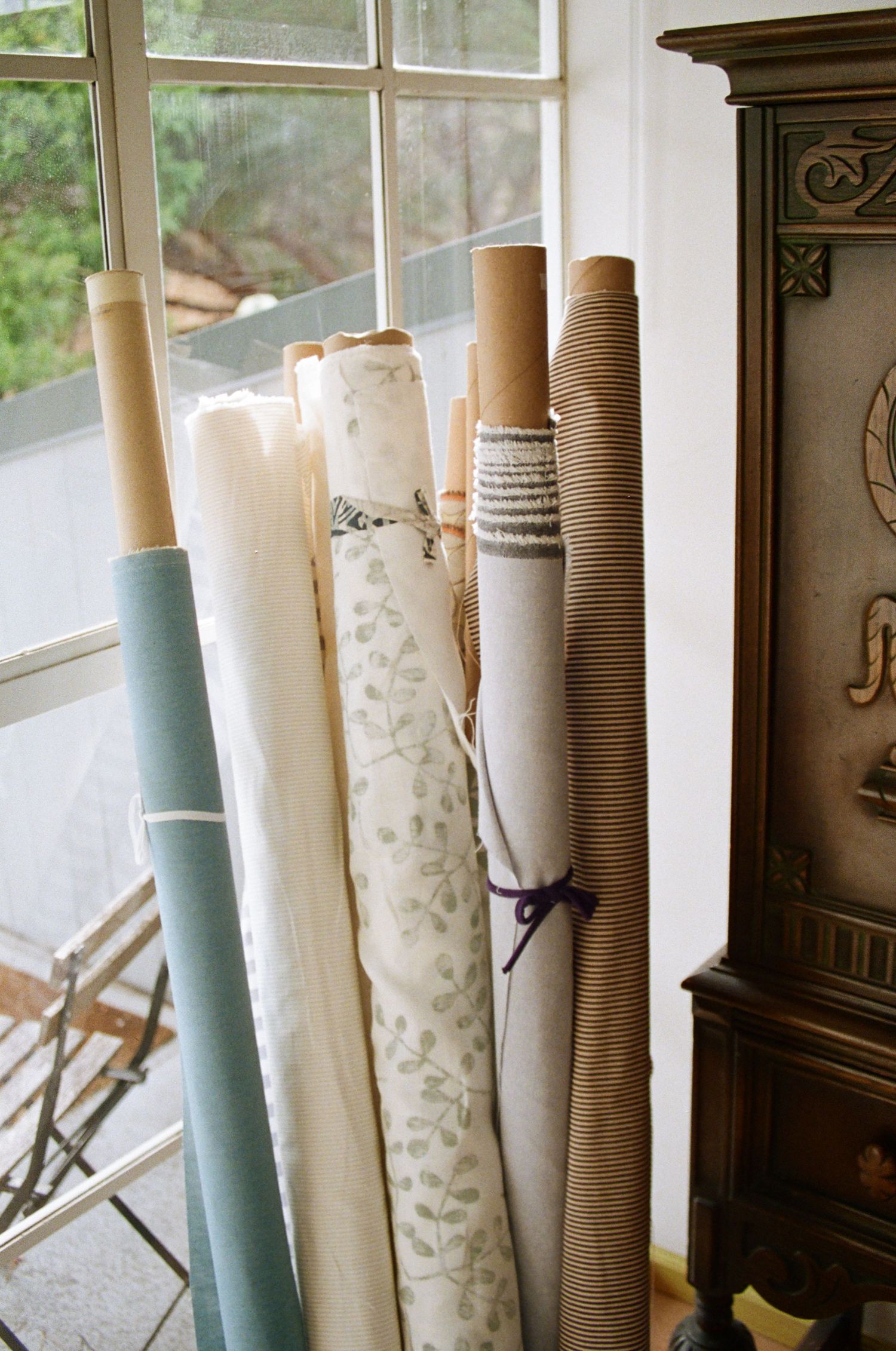 The image shows several rolls of fabric or wallpaper in various patterns and colors, leaning against a wooden cabinet in a window-lit room.