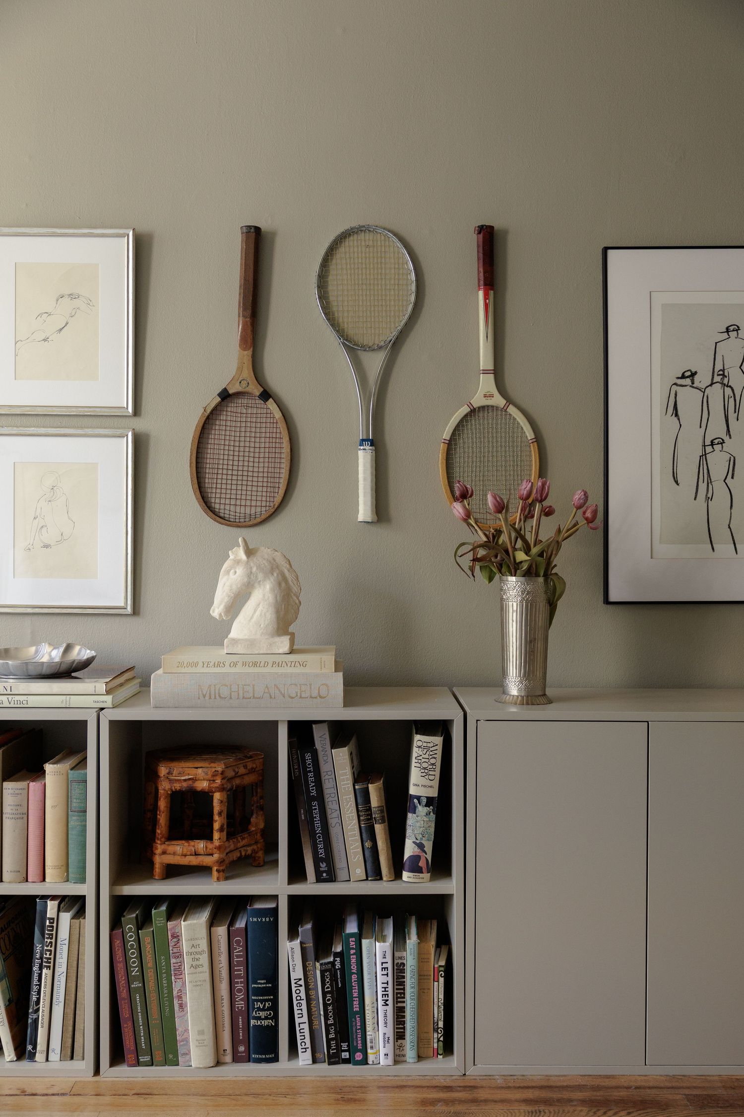 Vintage tennis rackets displayed on sage green wall above gray bookshelves with books, a horse sculpture, and pink flowers.