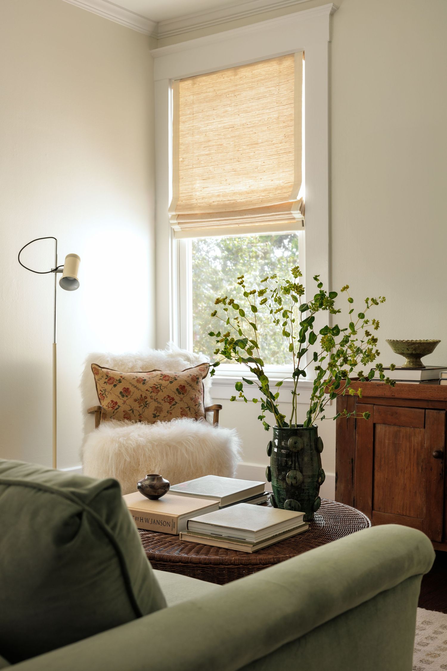 Cozy reading corner with bamboo shade, green velvet sofa, and potted plant by window.