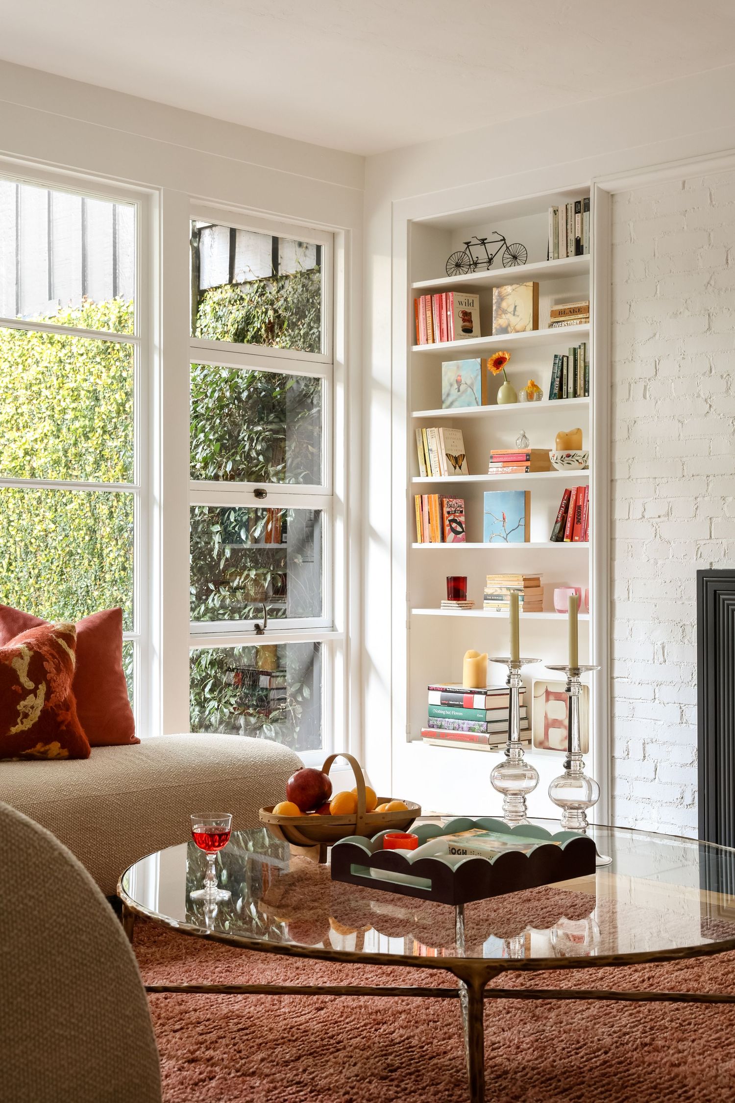 Bright living room with white bookshelf, glass coffee table, cozy sofa, and large windows overlooking greenery.