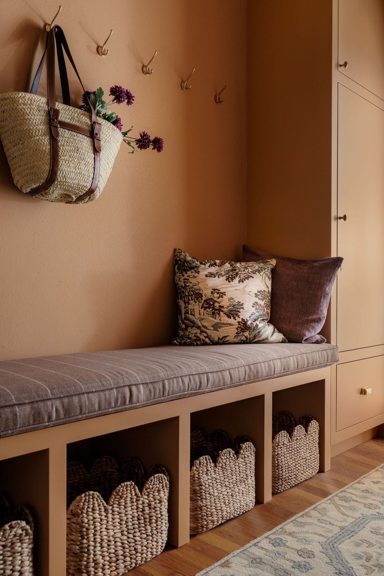 Warm terracotta mudroom with built-in bench, wicker storage baskets, decorative pillows, brass hooks, and a woven tote bag.