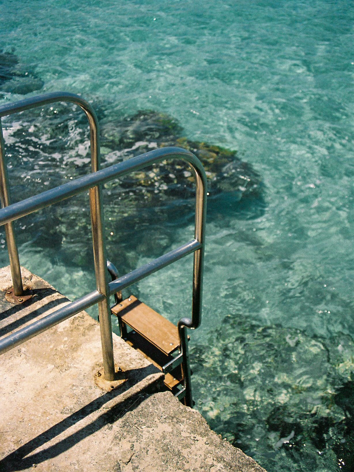 Metal handrail steps leading into clear turquoise ocean water from a rocky seaside platform.