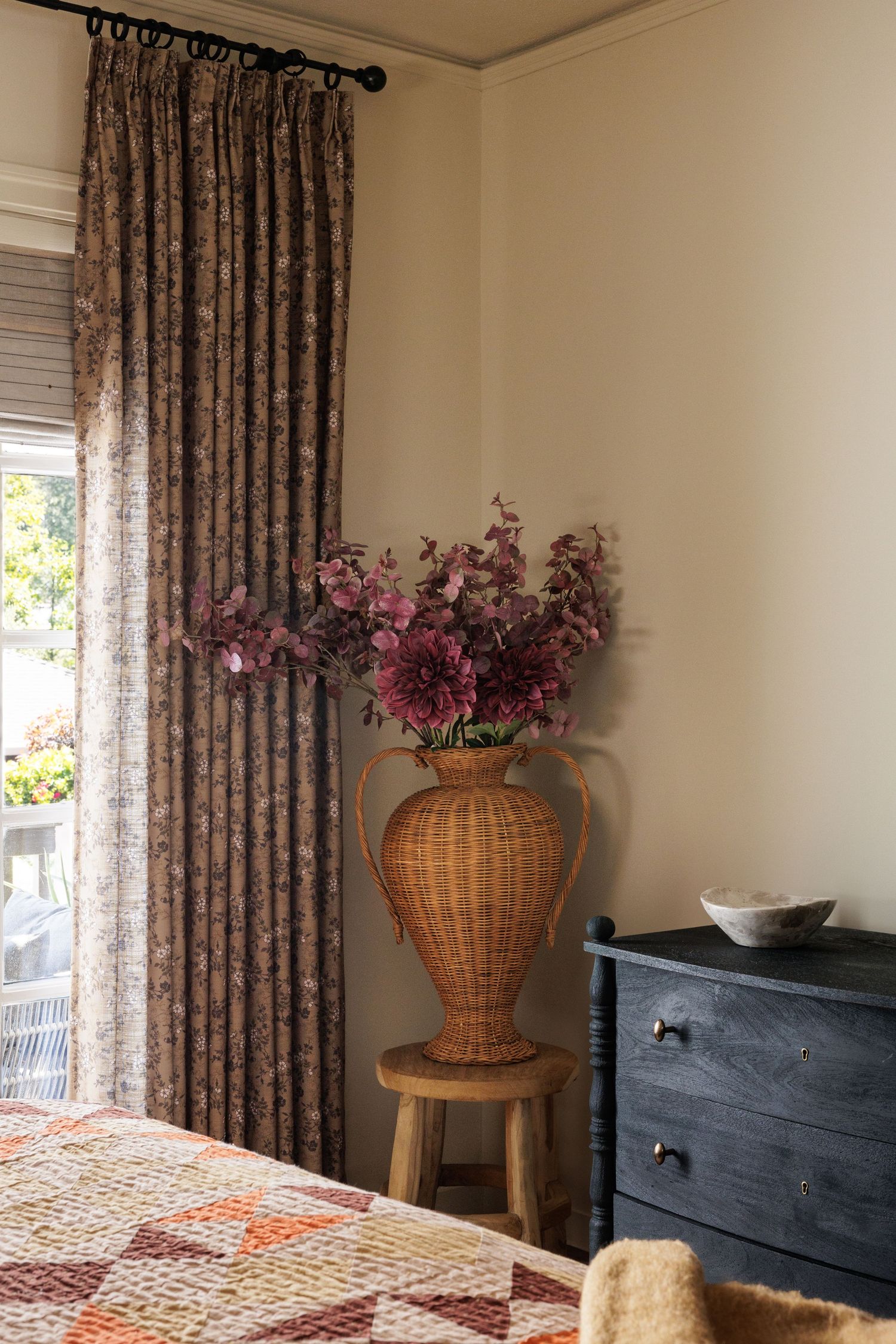 Bedroom corner with floral curtains, wicker chair, and pink flower arrangement.