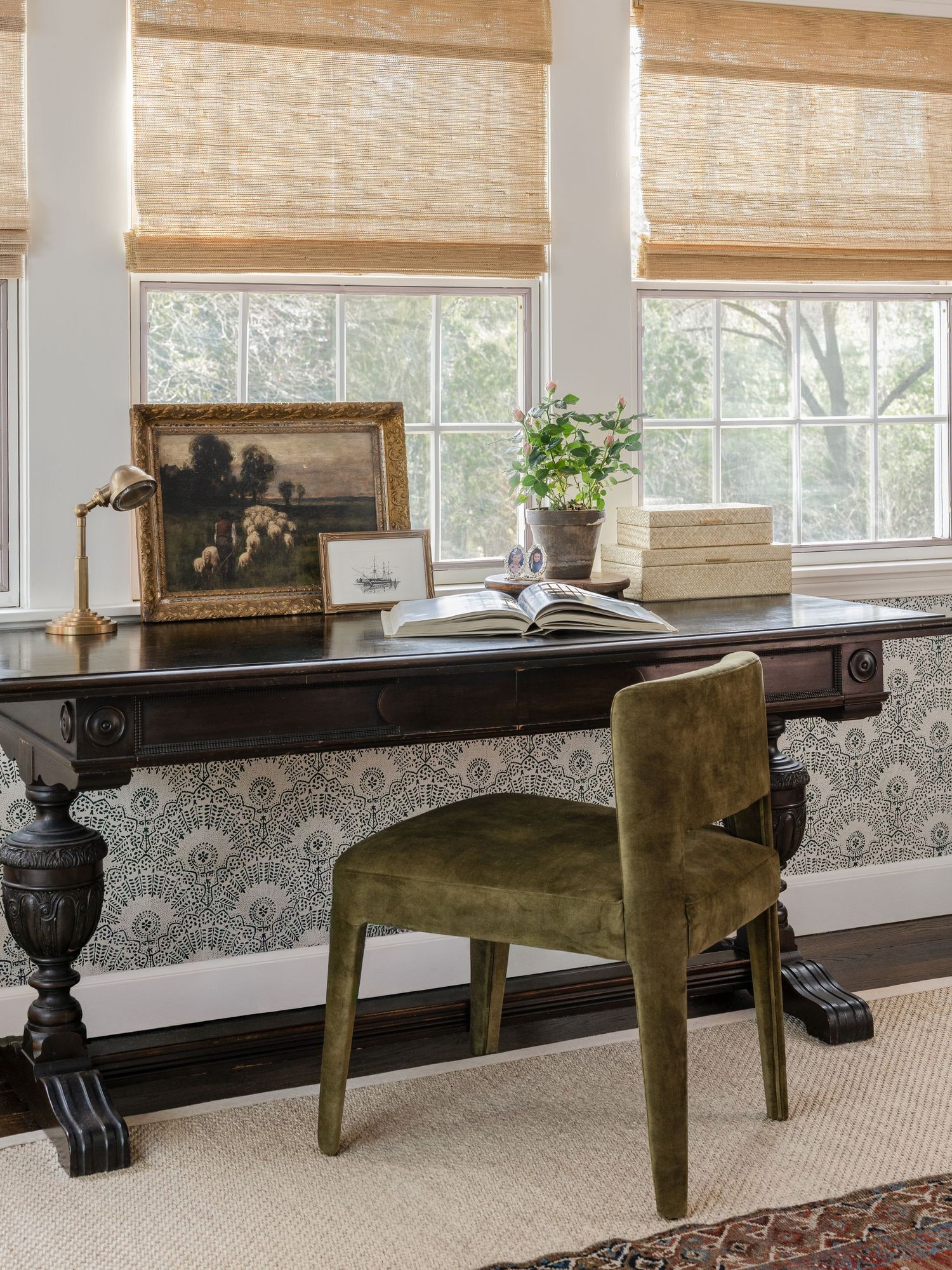 Elegant home office with dark wood trestle desk, green velvet chair, woven roman shades, and floral wallpaper backdrop.