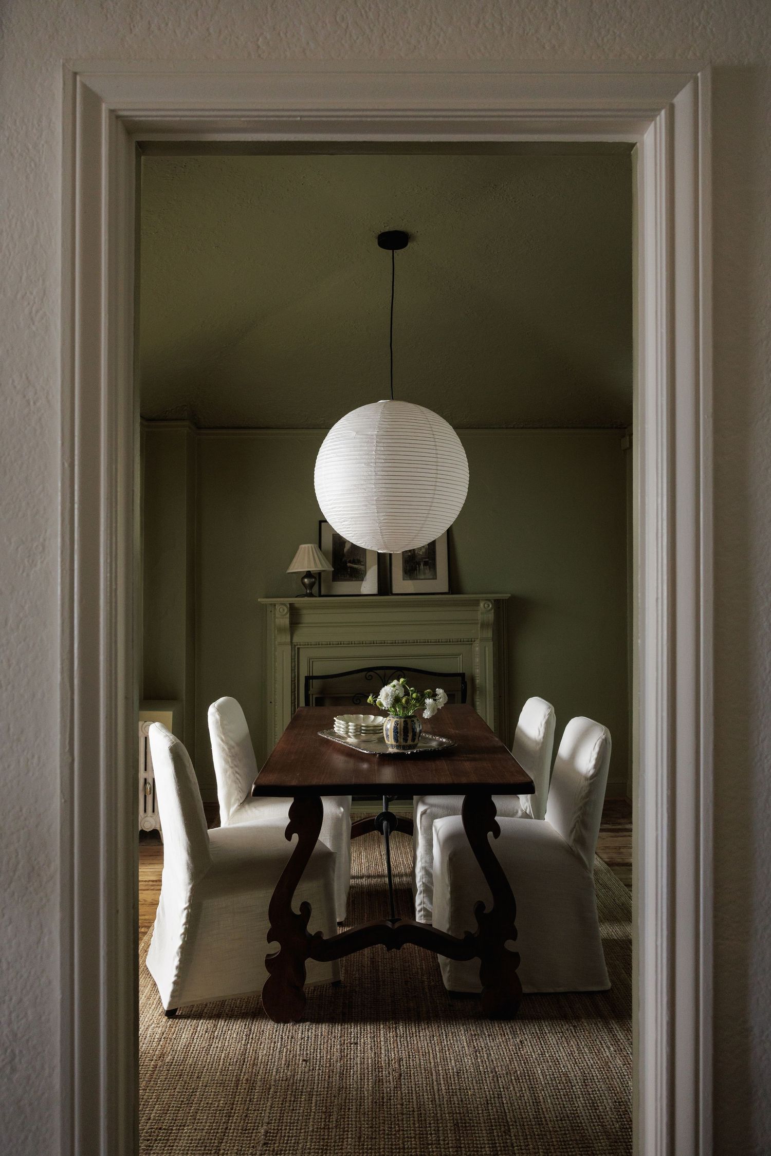 Elegant dining room with sage green walls, rustic wood table, white slipcovered chairs, and a globe pendant light.