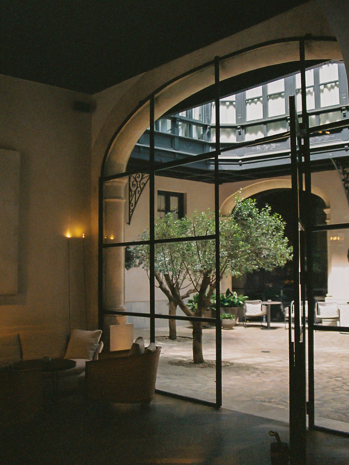 Elegant arched glass doors open to a sunlit courtyard with olive trees in a sophisticated hotel interior.