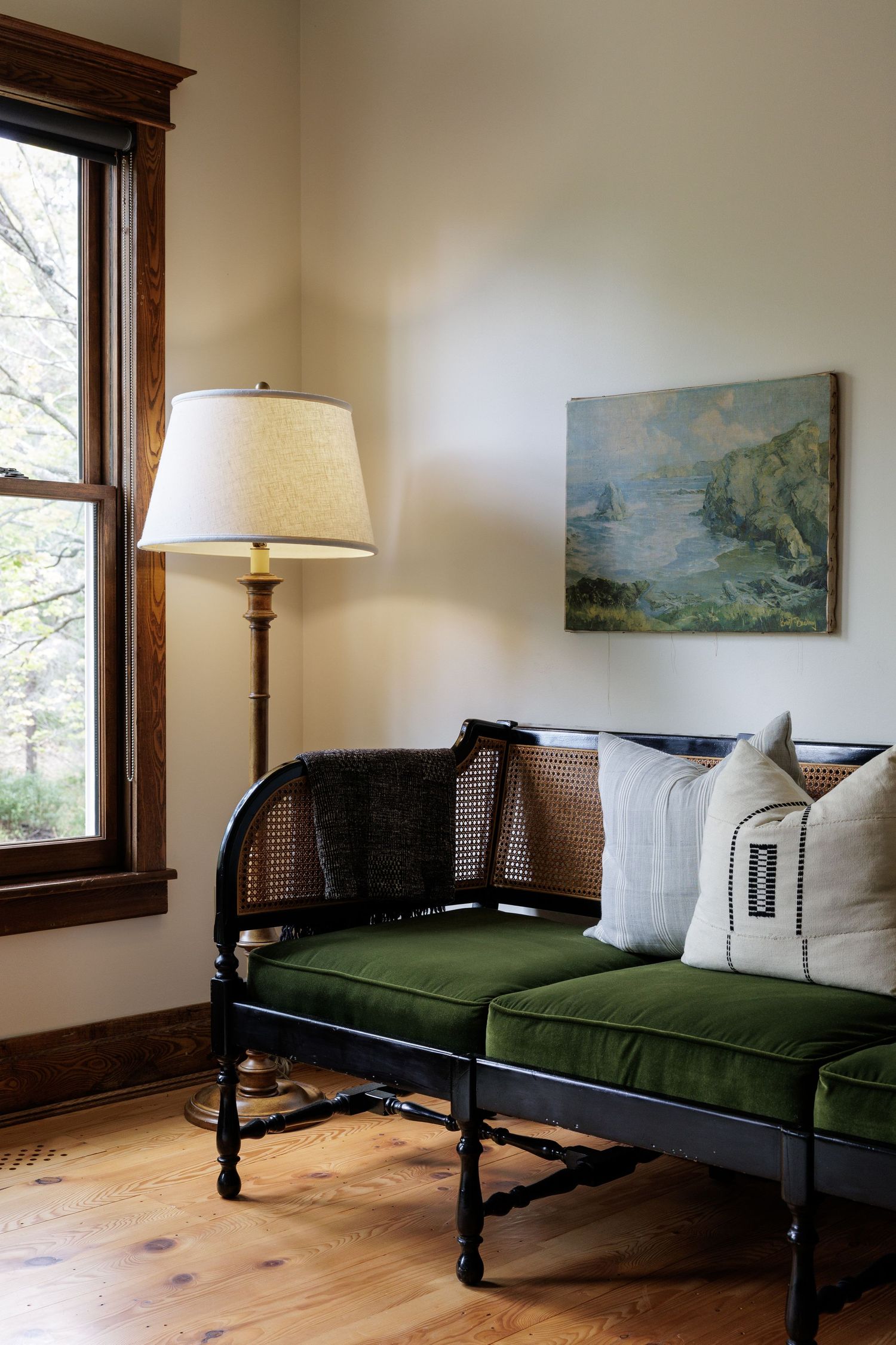 Living room with green cushioned daybed, landscape artwork, and wood-trimmed windows.