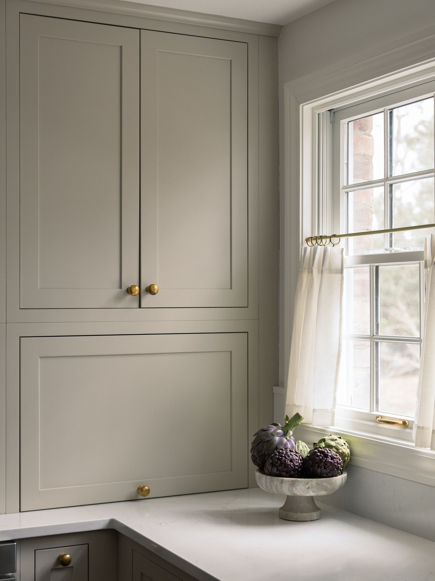 Elegant gray kitchen cabinets with brass knobs beside a white window with sheer curtains and a marble countertop.