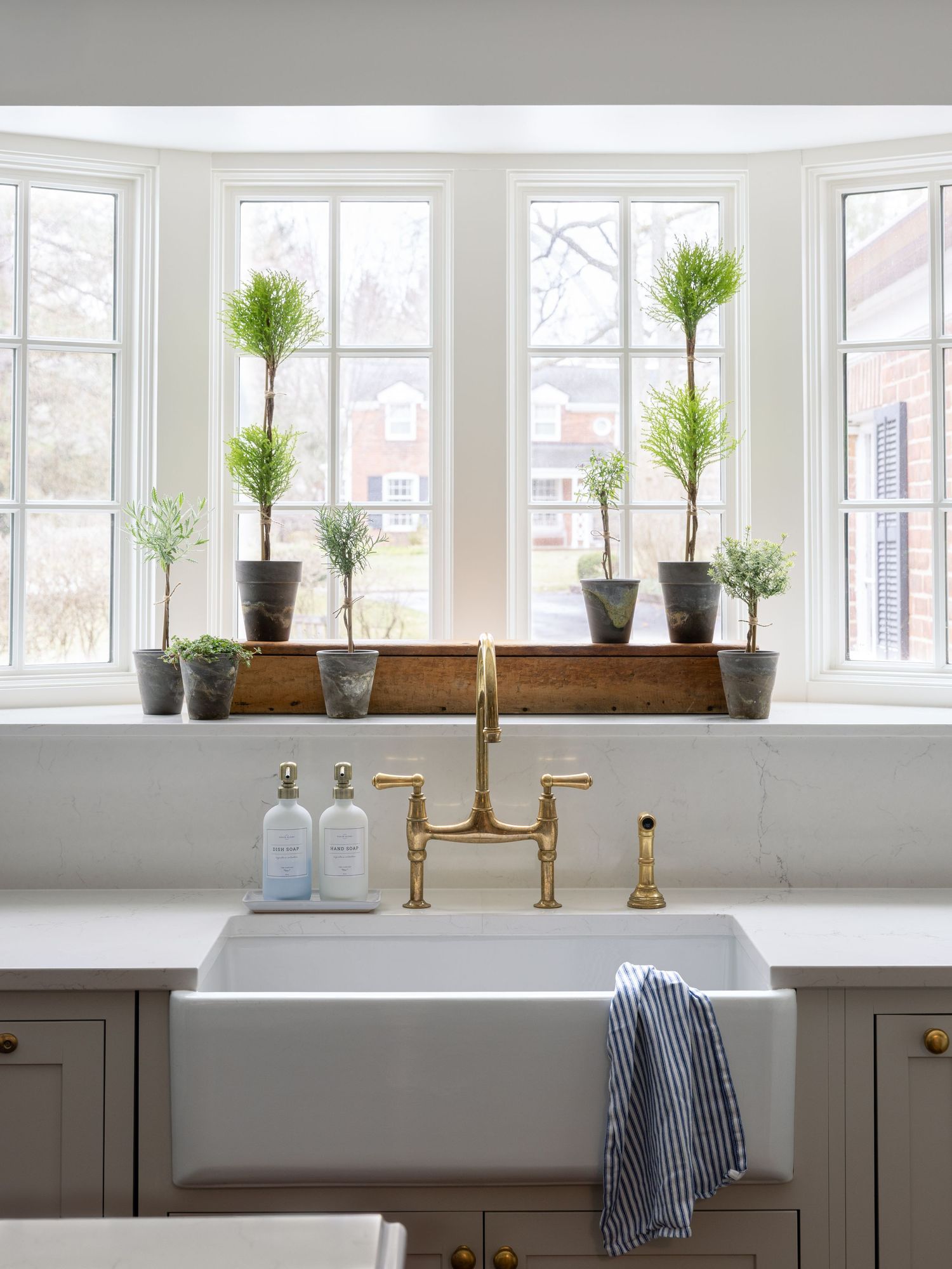 White farmhouse sink with brass faucet, gray cabinets, and potted topiaries on a bay window sill in a bright kitchen.