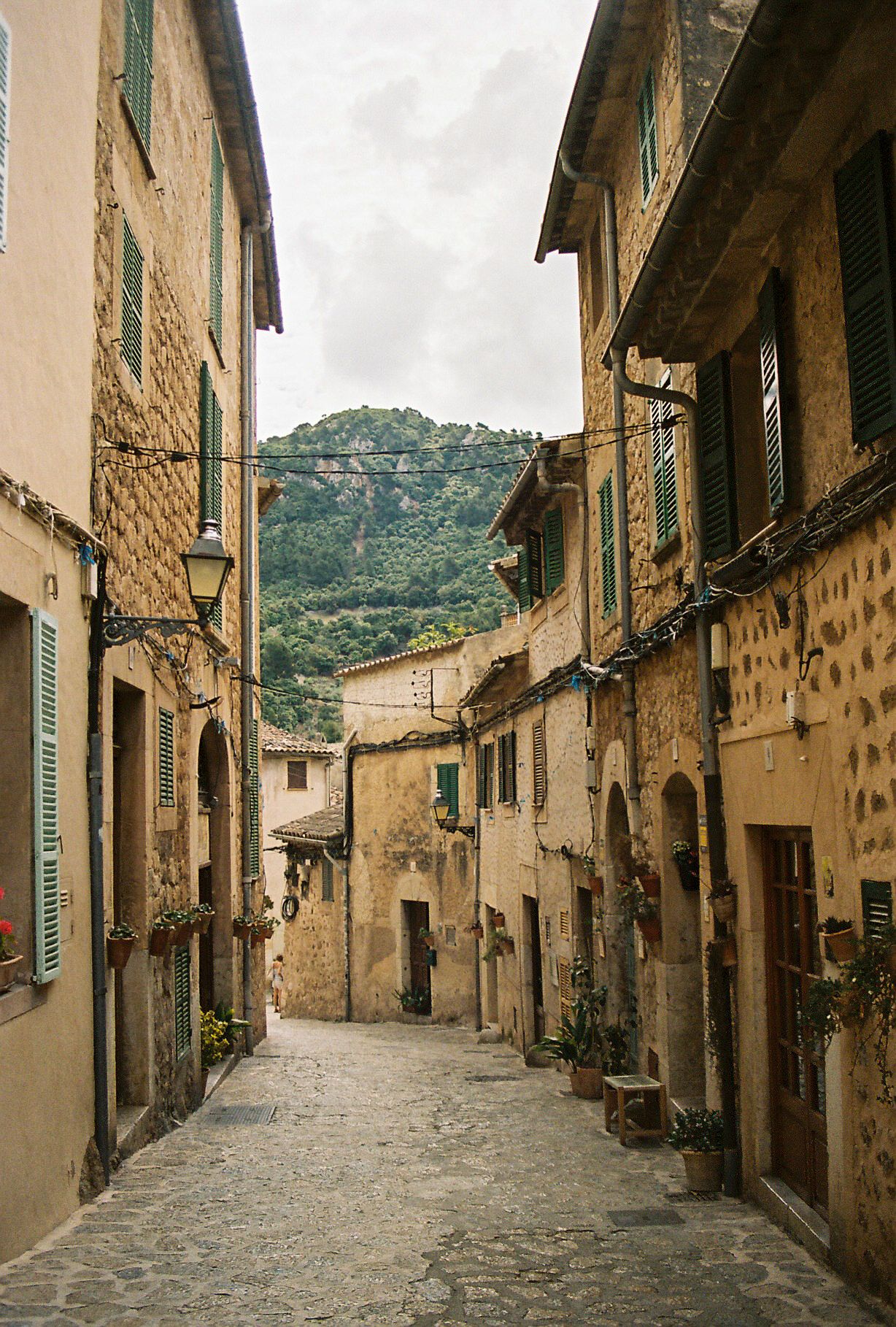 Narrow cobblestone alley lined with ancient stone buildings in a charming Mediterranean village with green hills.
