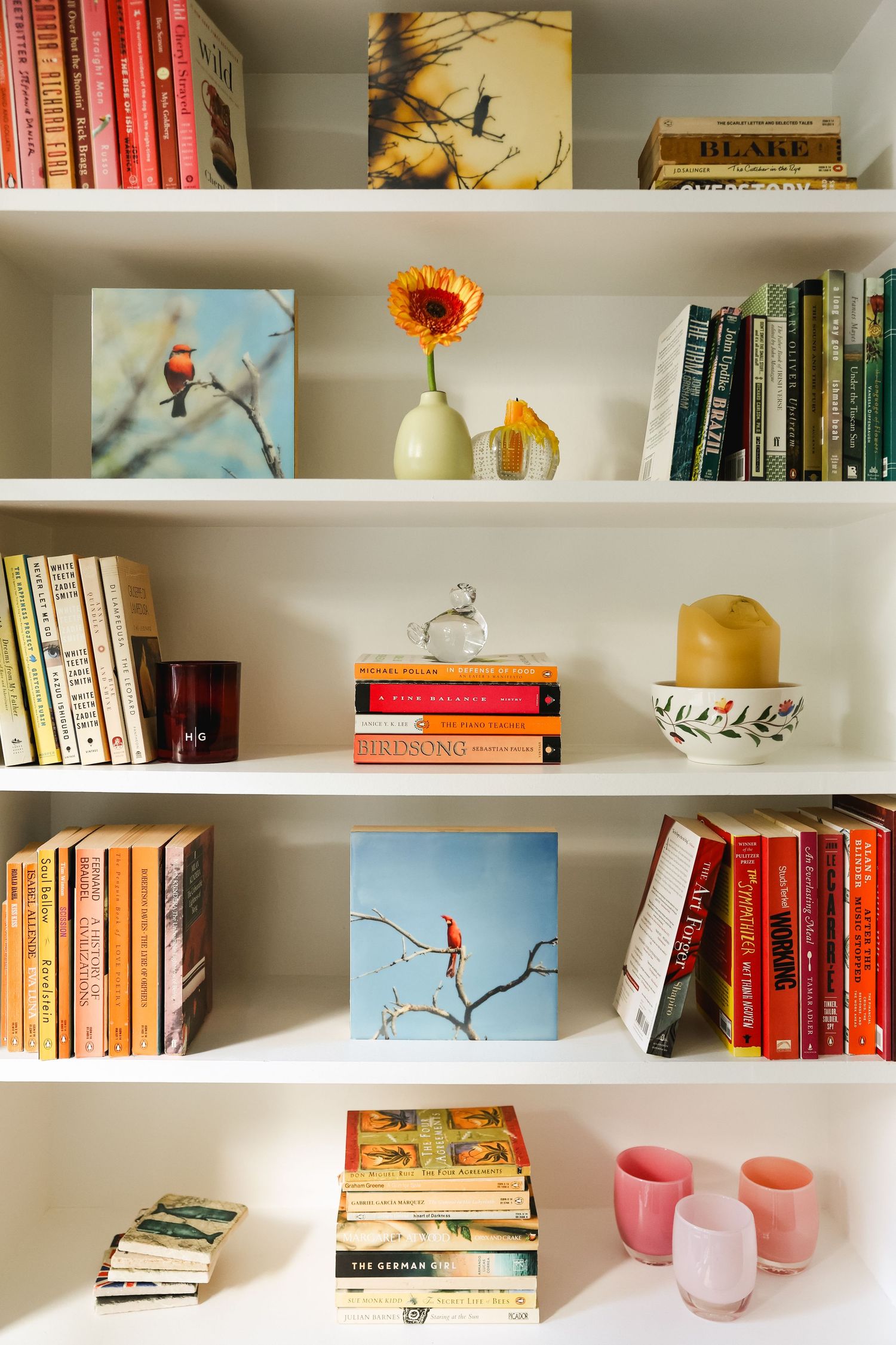 White built-in bookshelves styled with colorful books, bird artwork, a sunflower vase, candles, and pink ceramic cups.