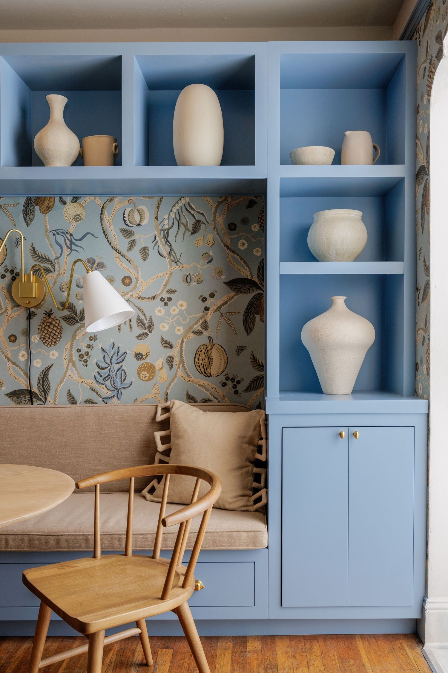 Blue built-in shelving with ceramic vases, floral wallpaper, cozy bench seating, and a wooden chair in a stylish nook.