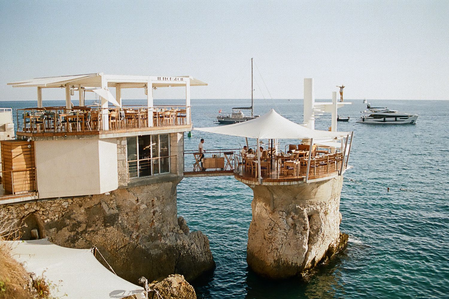 Oceanfront restaurant built into rocky cliffs overlooking blue Mediterranean waters.
