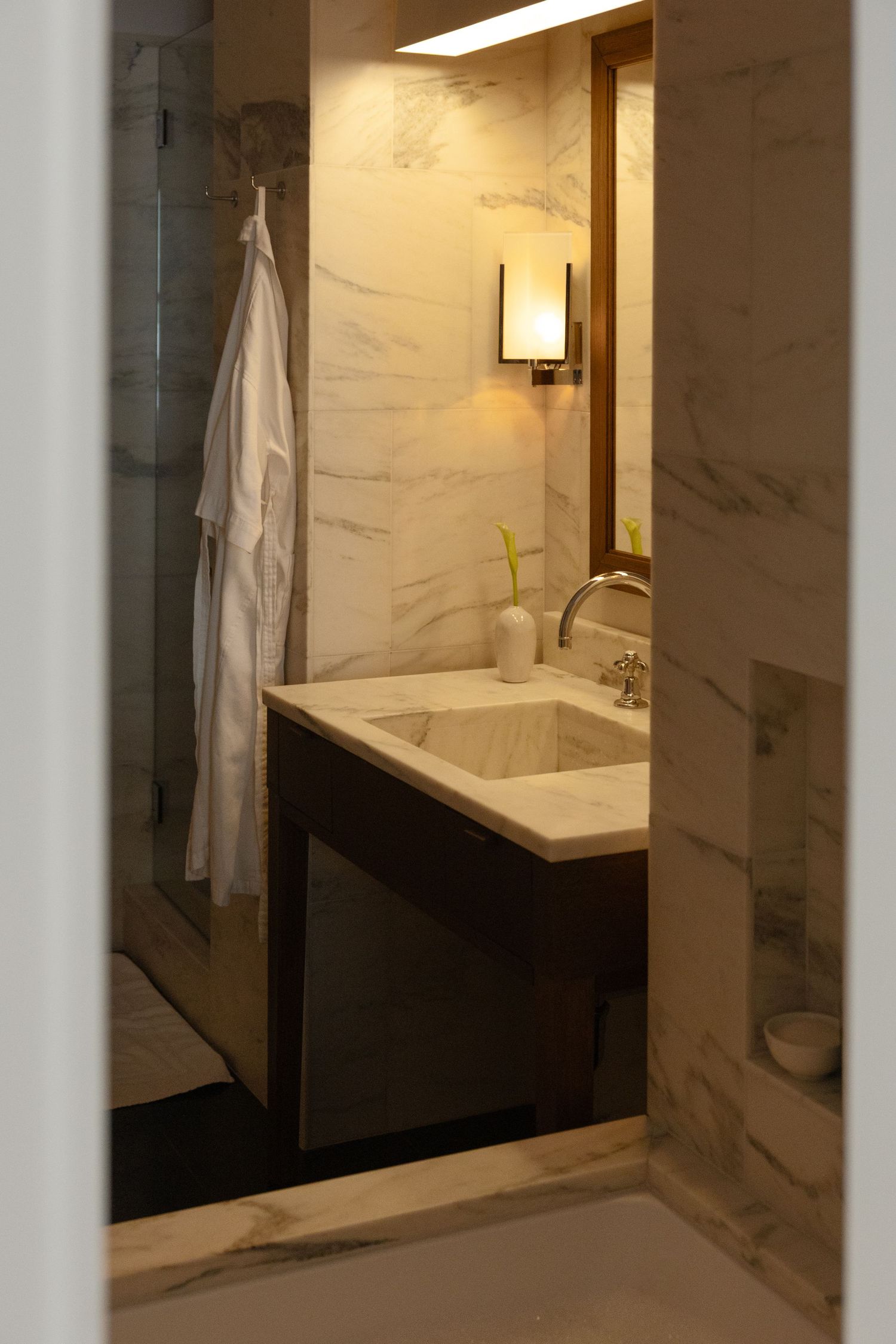 Elegant marble bathroom with dark vanity, wall sconce, and white robe hook.