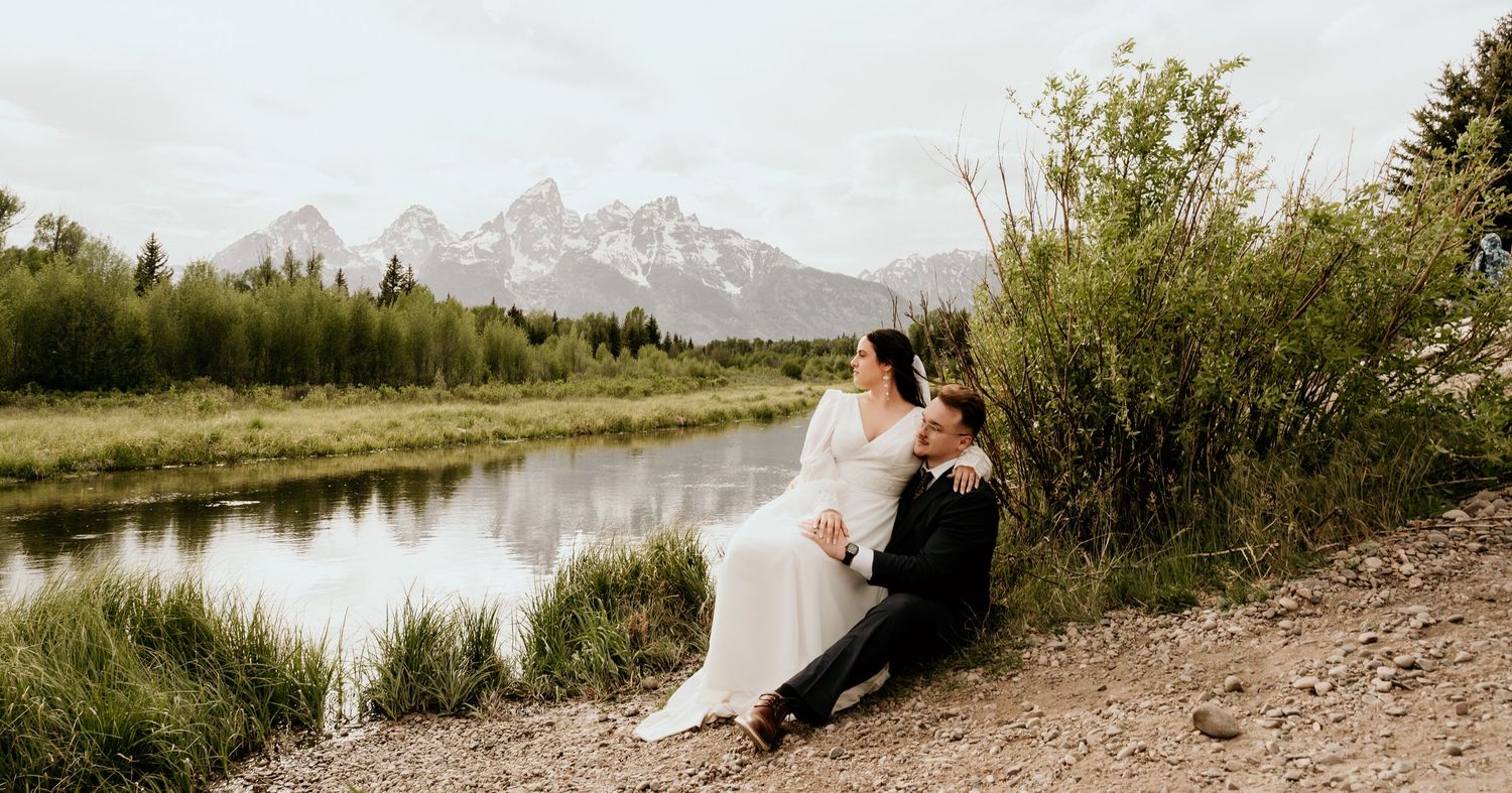Bride and groom sitting together on rocky shore beside a river with Grand Teton mountains in the background.