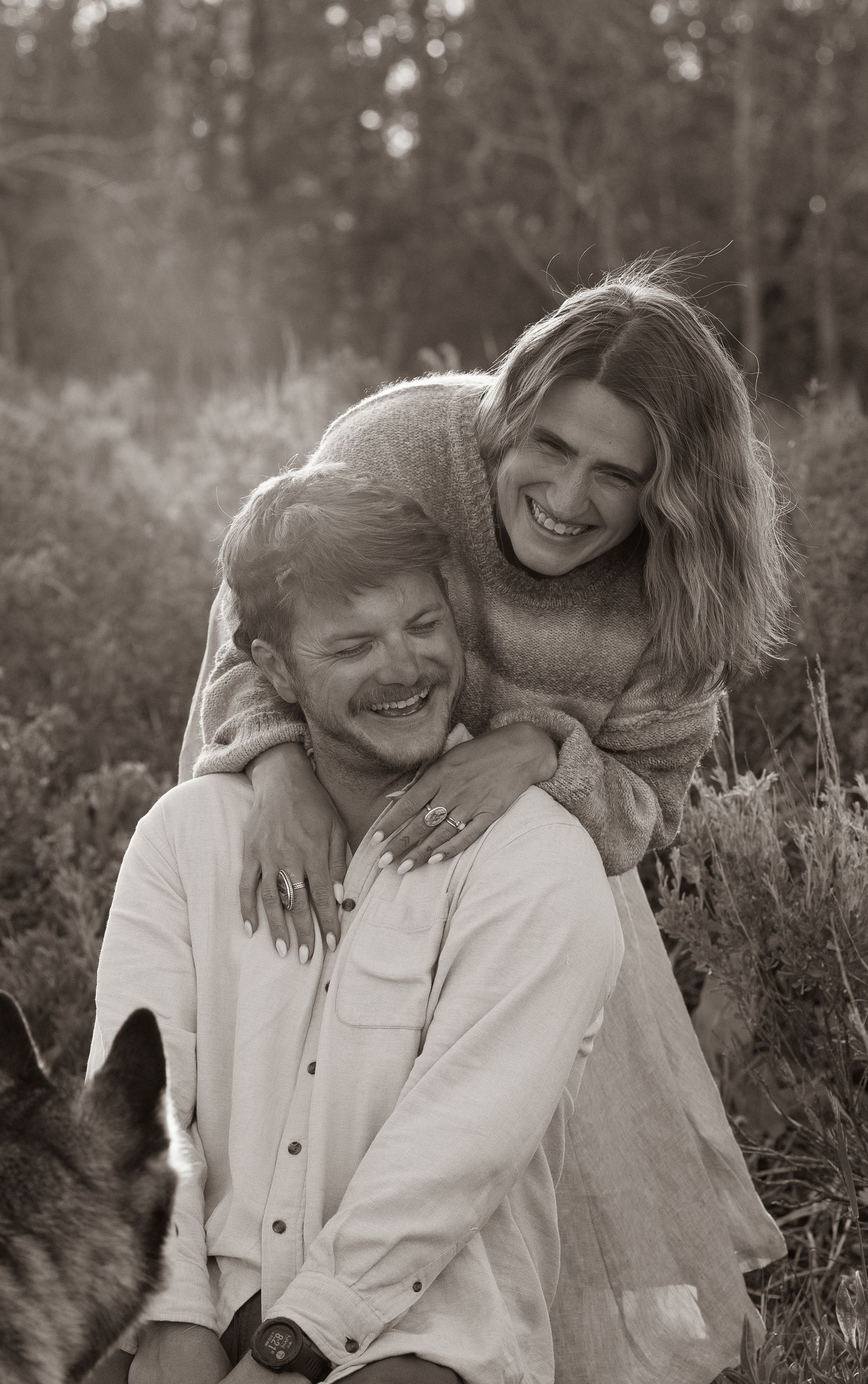 Laughing couple embracing outdoors in sepia tone with German Shepherd dog visible in foreground.