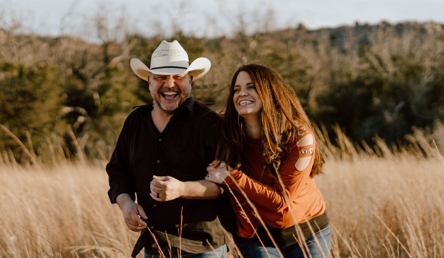 Couple poses playfully in tall grass field during sunset.