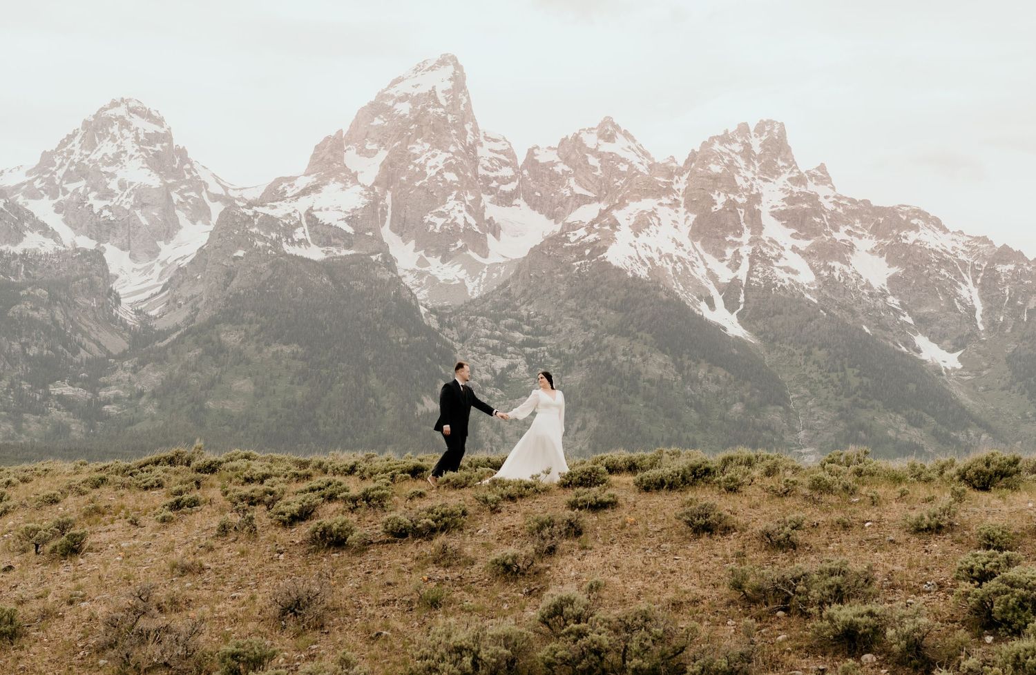 Bride and groom hold hands running on a hillside with snow-capped Grand Teton mountains in the background.