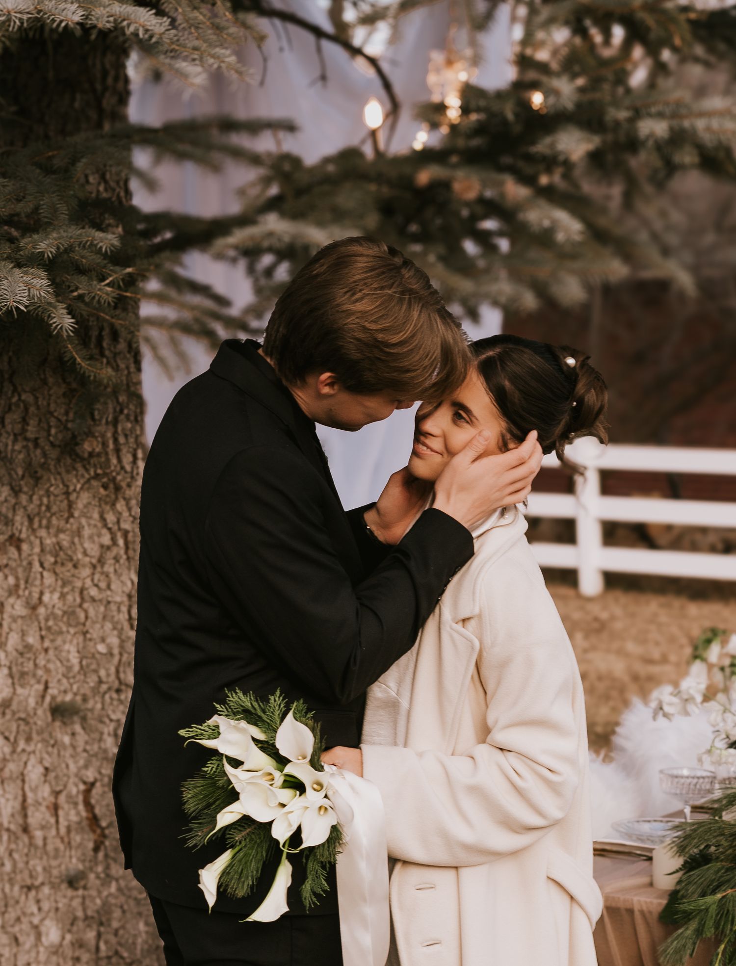 A couple shares an intimate moment outdoors near a white fence while holding a bouquet of white lilies.