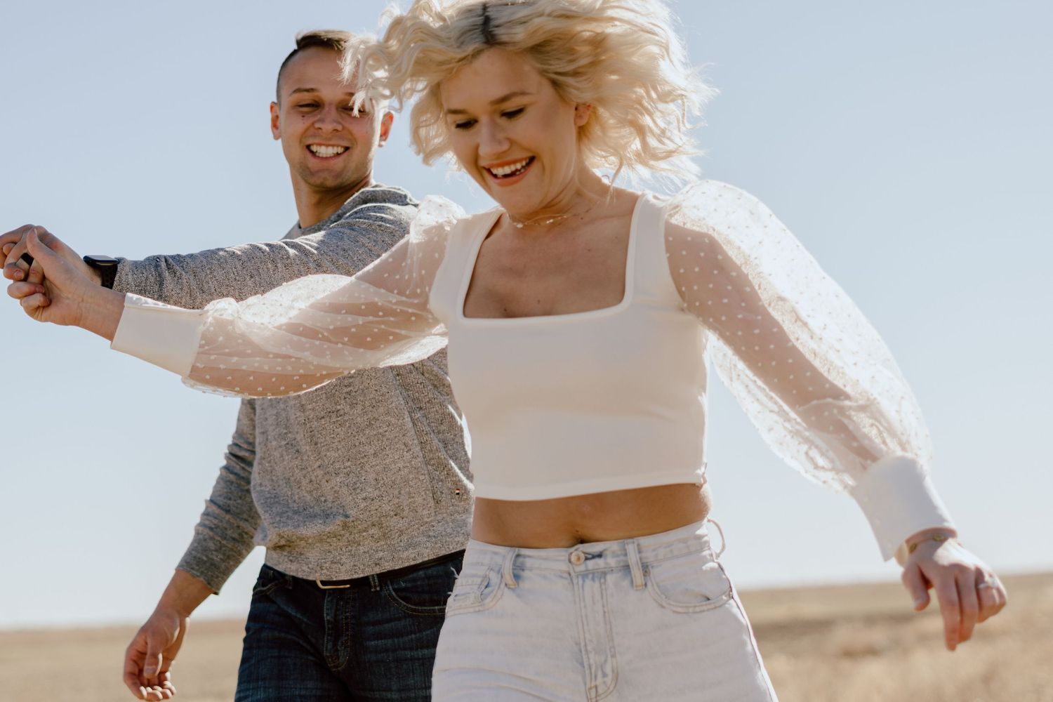 Playful couple dances together in casual outfits against bright sky background.