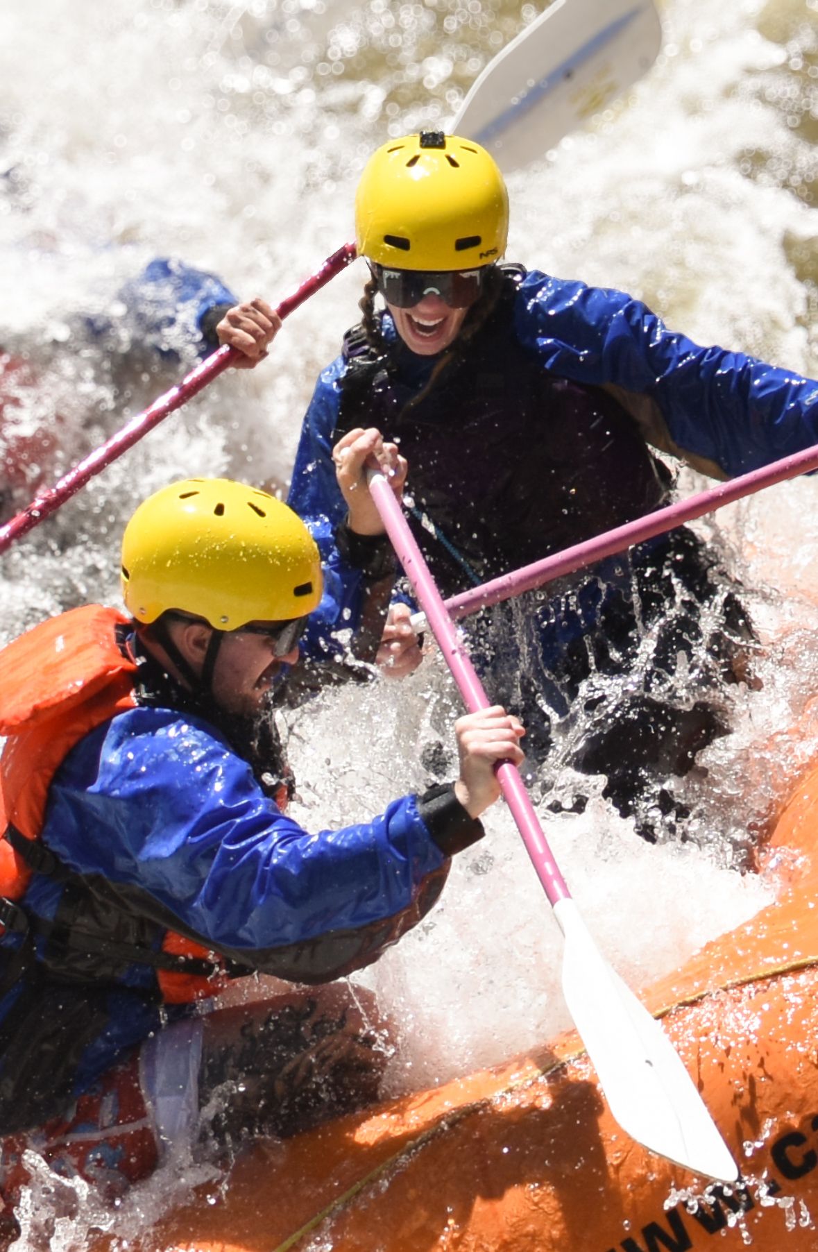 White water rafters navigate rapids in orange raft wearing blue gear and yellow helmets.