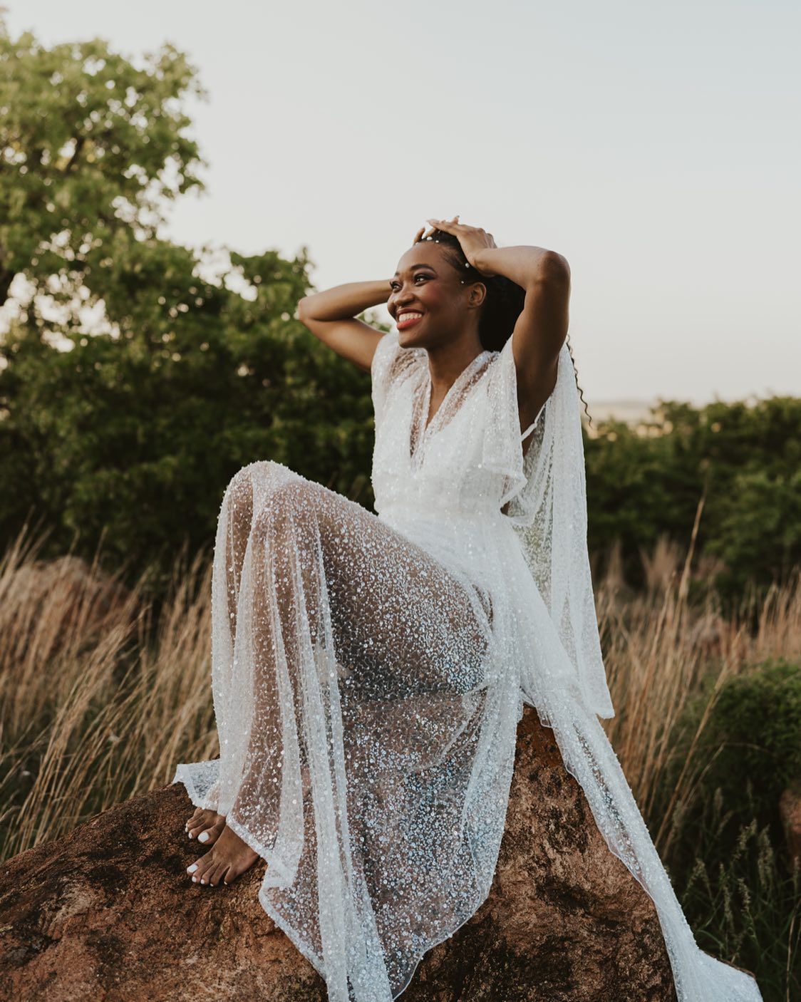 A flowing white lace dress photographed in golden light against tall grass and trees.