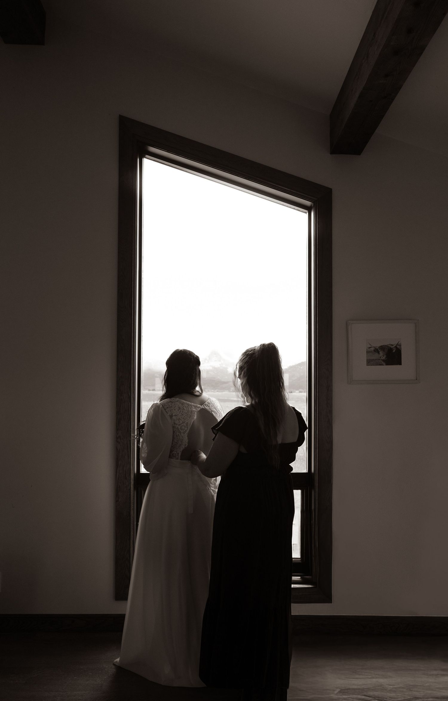 Bride and bridesmaid silhouetted in a large window in sepia tone.