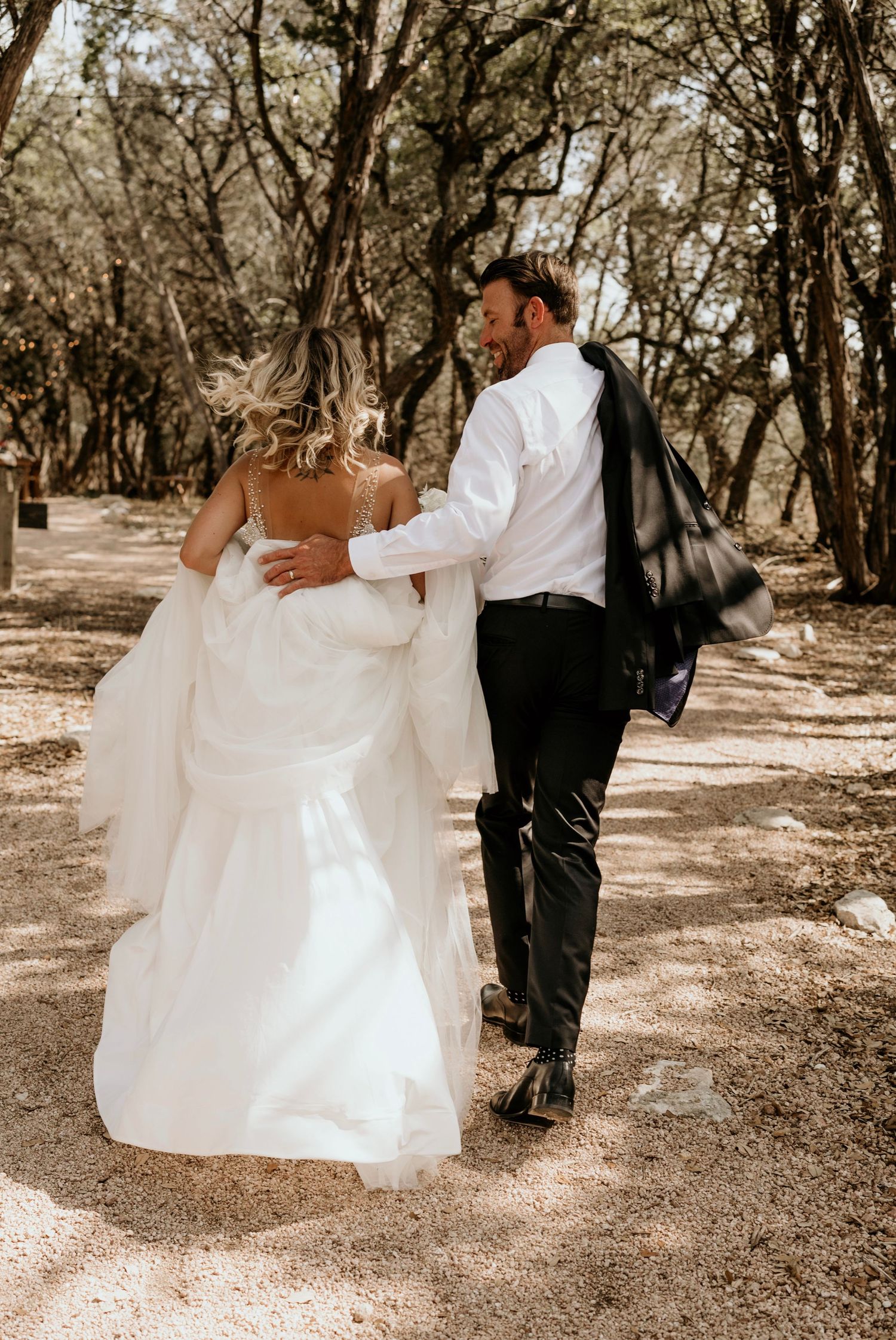 A couple in wedding attire walks hand in hand down a tree-lined path in warm sepia tones.
