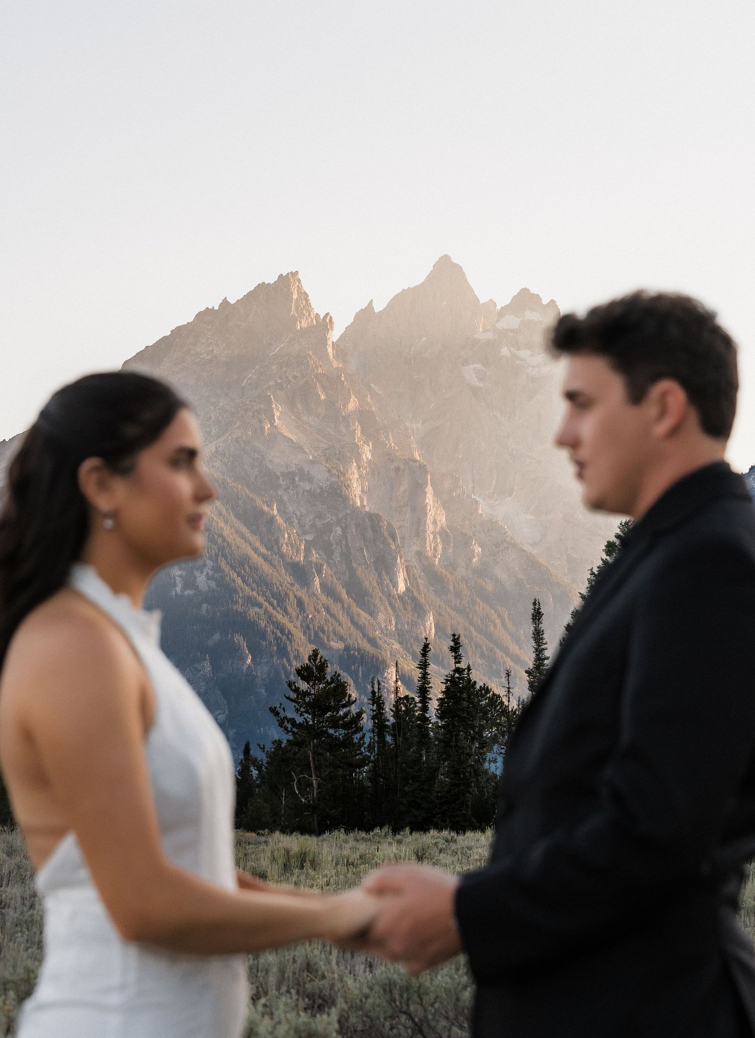 Couple holds hands during mountain elopement ceremony with Grand Teton peaks glowing at golden hour in background.