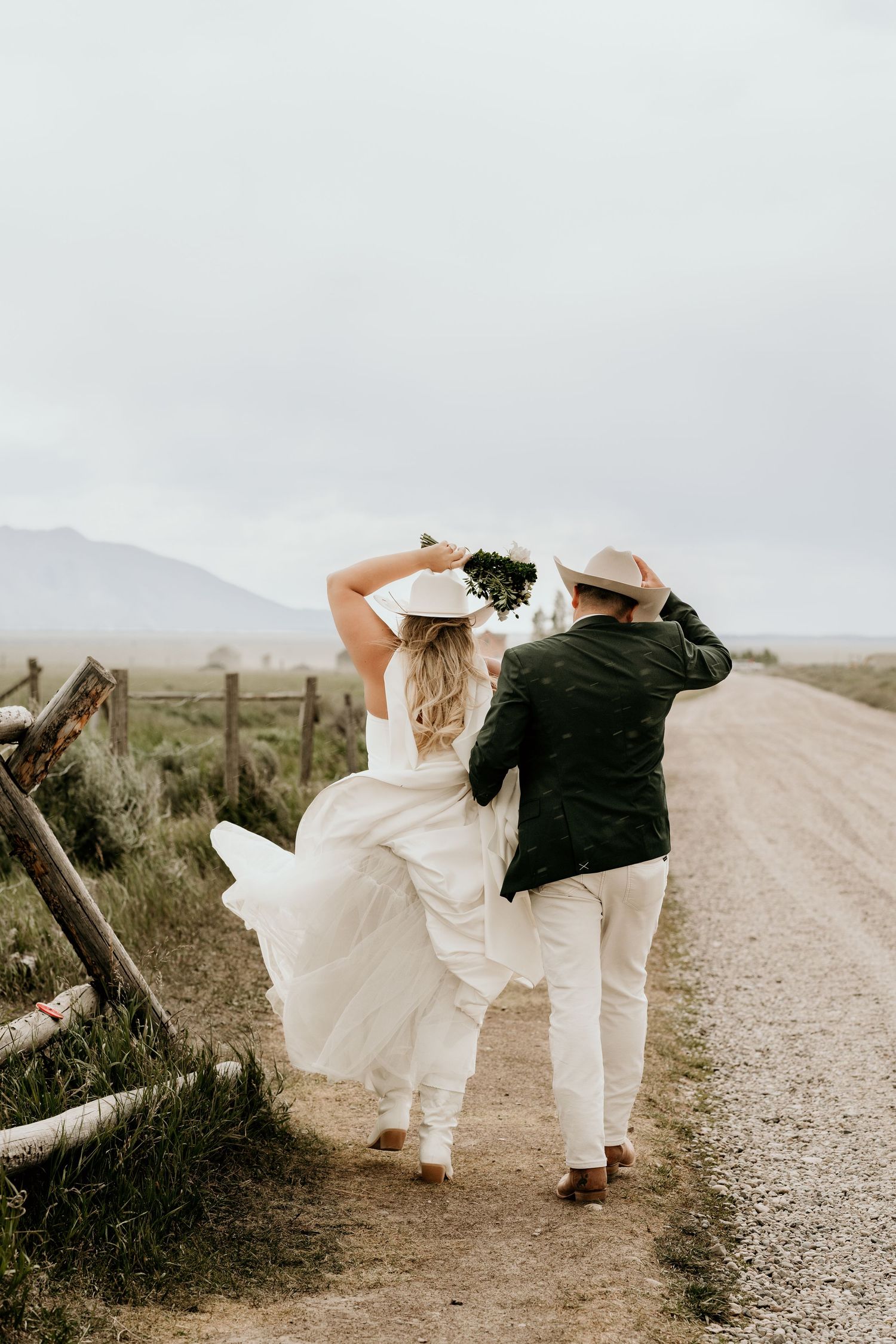 A bride and groom walk hand-in-hand down a rustic dirt road, holding their hats in the wind on their wedding day.