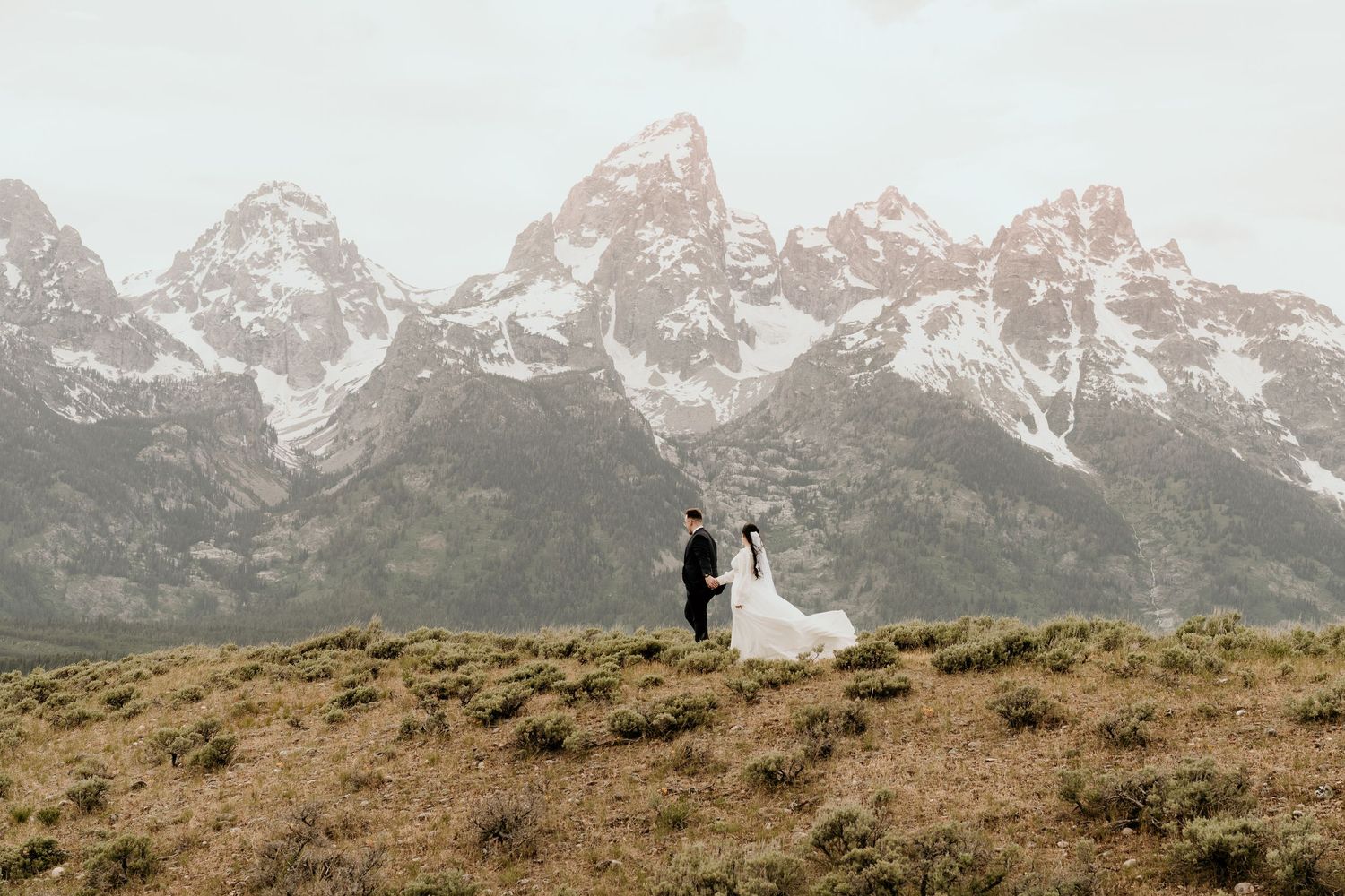 Bride and groom hold hands on a grassy hill with snow-capped Grand Teton mountains in the background.