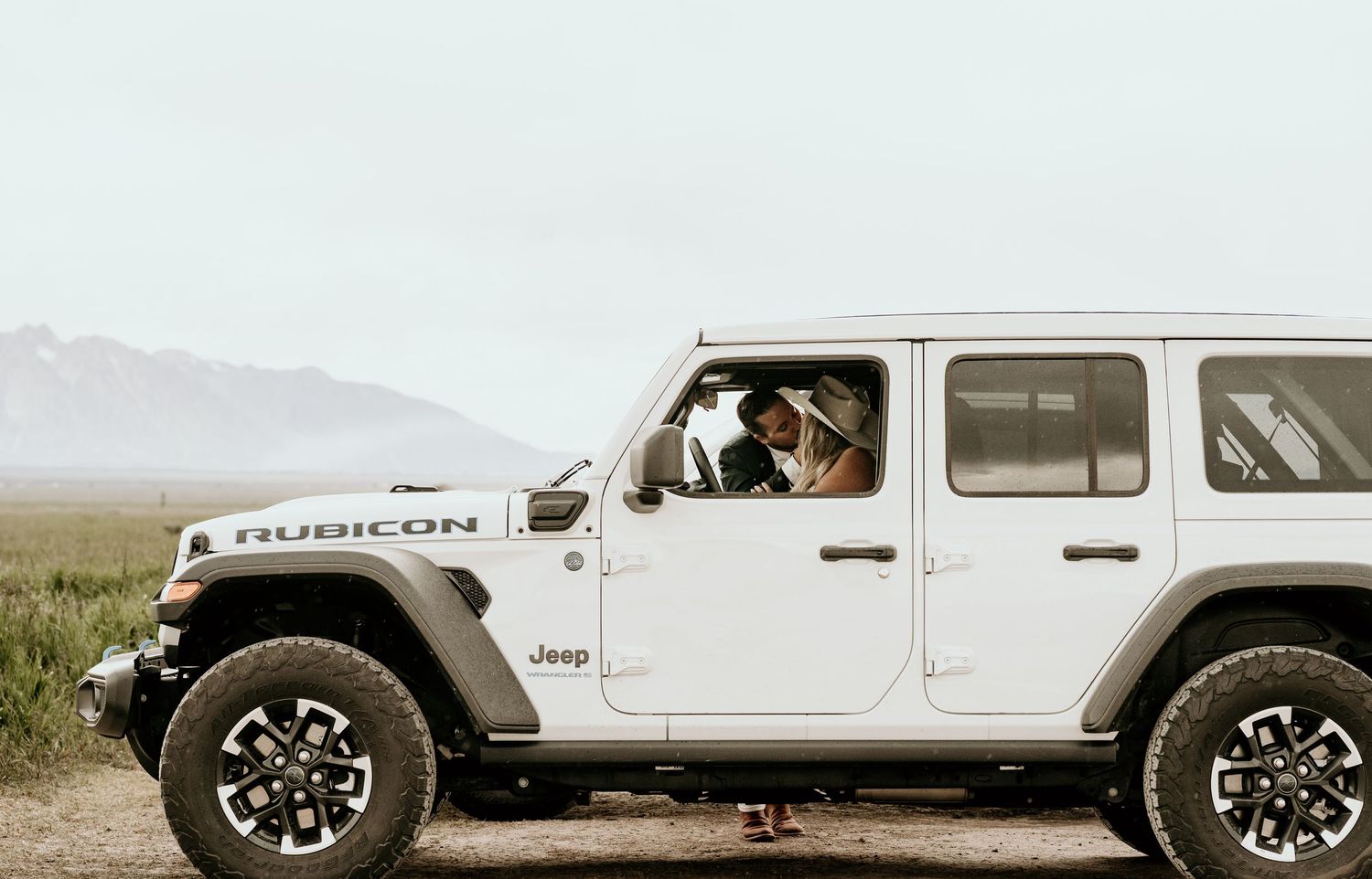 Couple in a white Jeep Wrangler Rubicon driving through open countryside with mountains in background.