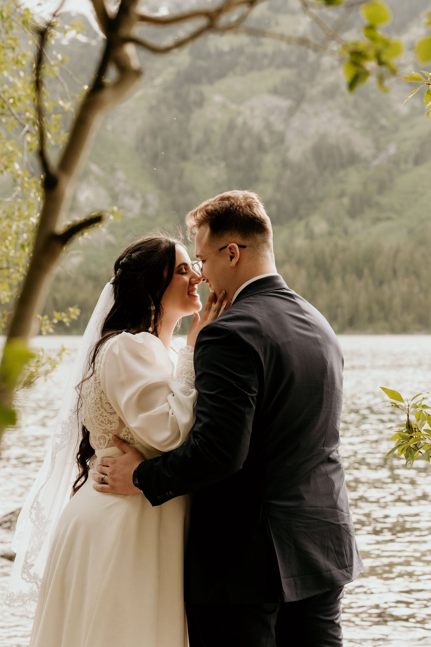 A bride and groom share a romantic moment by a mountain lake, with the bride in a white gown and veil.