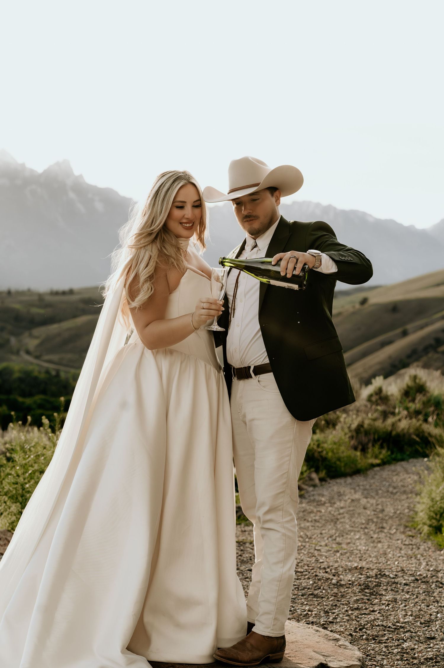 A bride and groom pour champagne outdoors with mountain views, she in a white gown, he in a suit and cowboy hat.