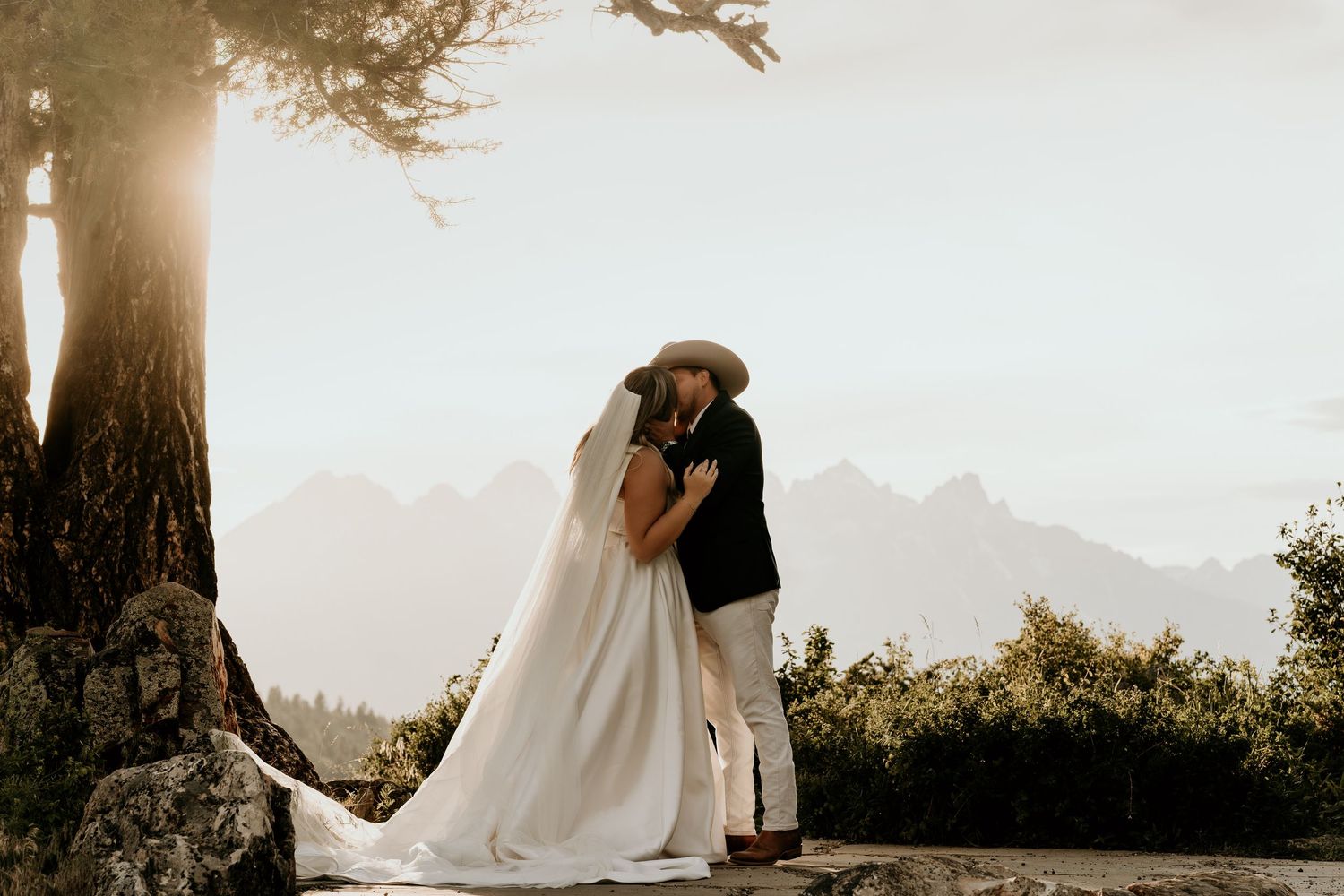 Bride and groom share a romantic kiss at mountain overlook, sun-drenched backdrop with misty peaks and rustic tree.