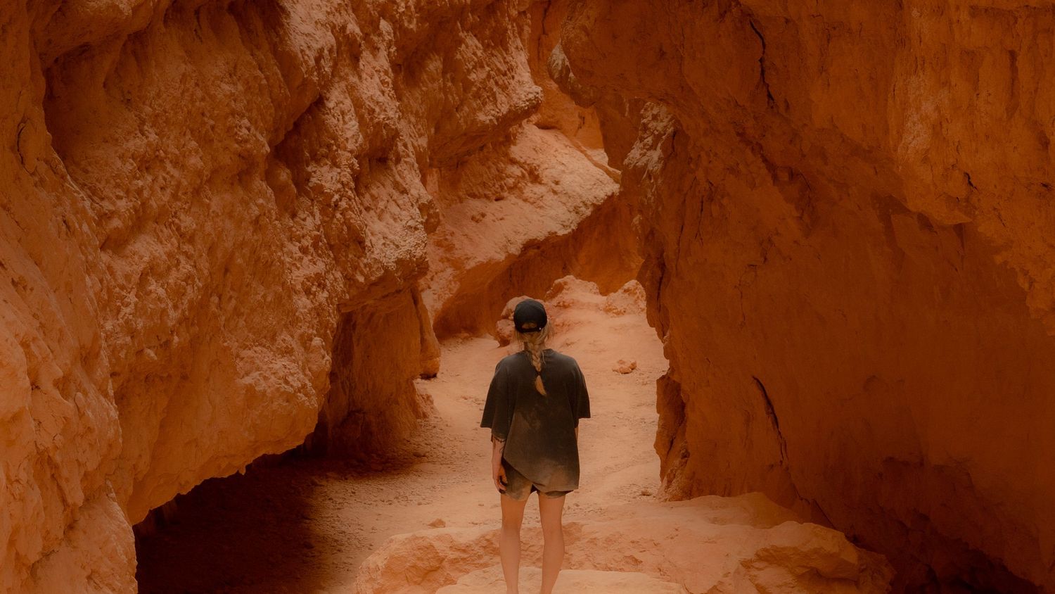 A person walks through a narrow sandstone canyon with towering orange rock walls glowing in filtered sunlight.