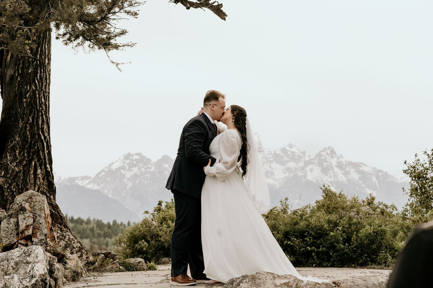 Bride and groom share a romantic kiss with snow-capped mountains and evergreen trees in the background.