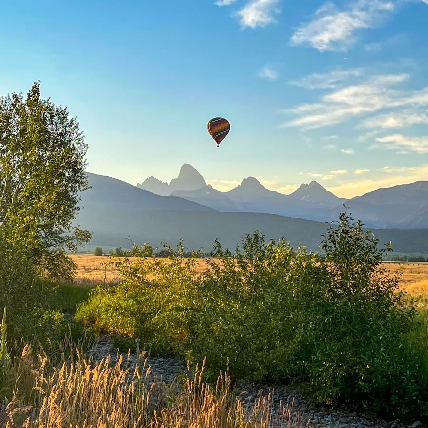 Hot air balloon floats above mountain landscape with Teton peaks silhouetted against sunset sky in Grand Teton National Park.