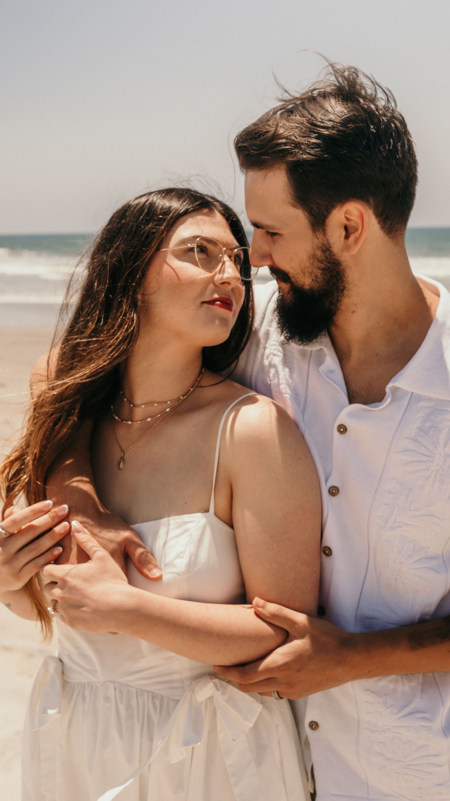 Romantic beach photoshoot of a couple in white clothing by the ocean.