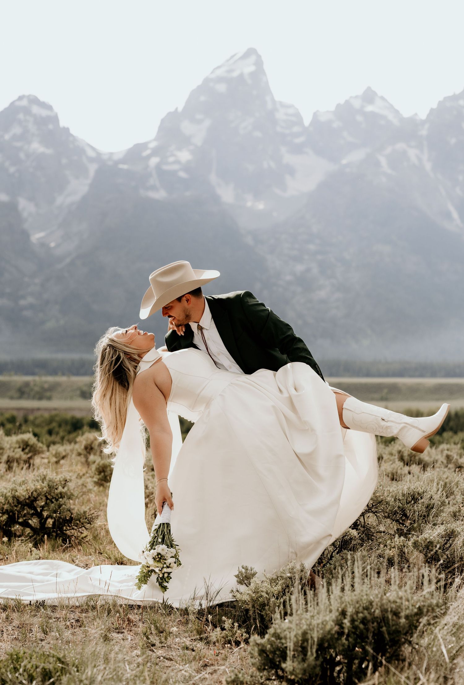 Groom lifts bride in cowboy hat and boots with Grand Teton mountains in background.