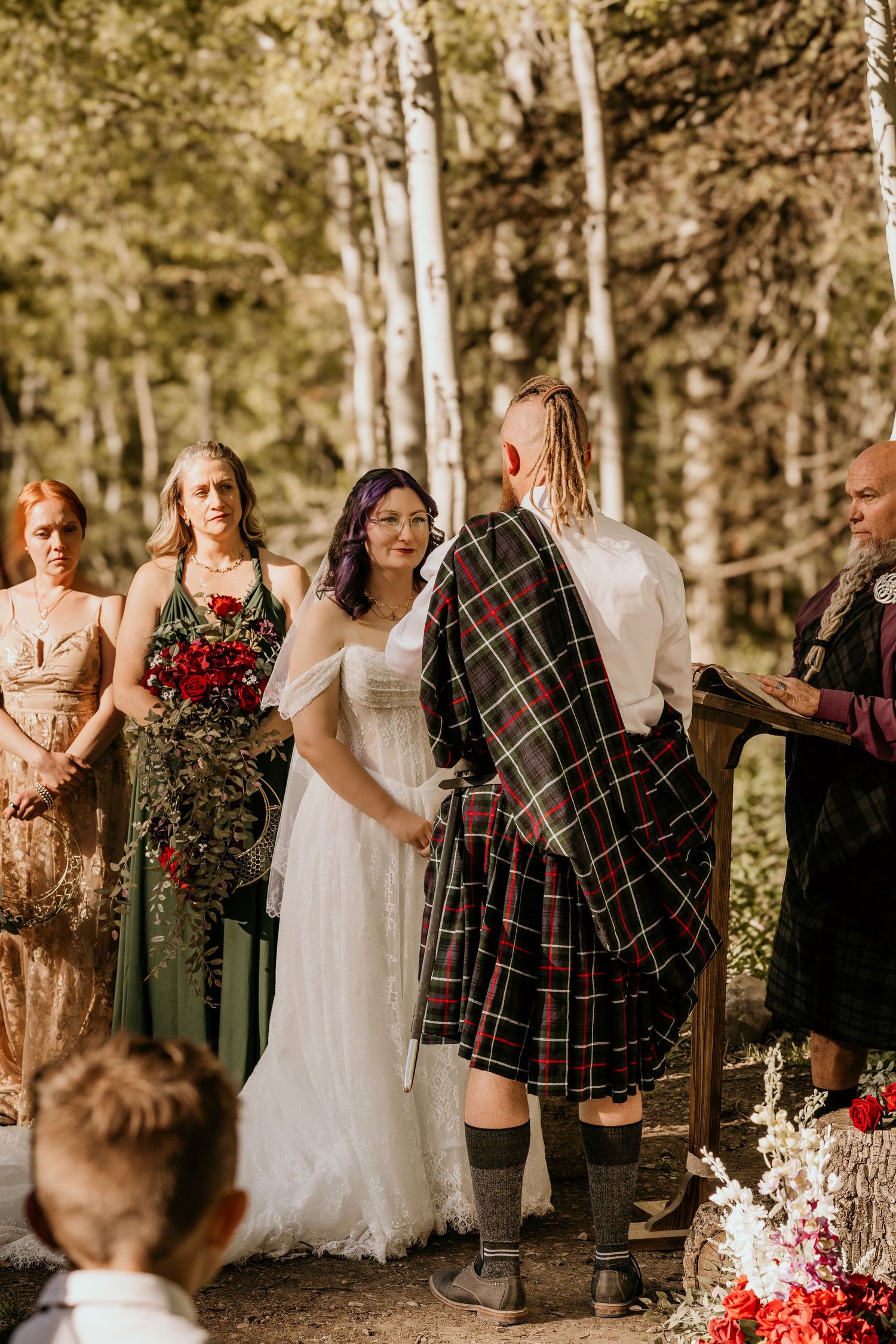 Outdoor forest wedding ceremony with bride in white dress and groom in tartan kilt, surrounded by bridesmaids holding red bouquets.