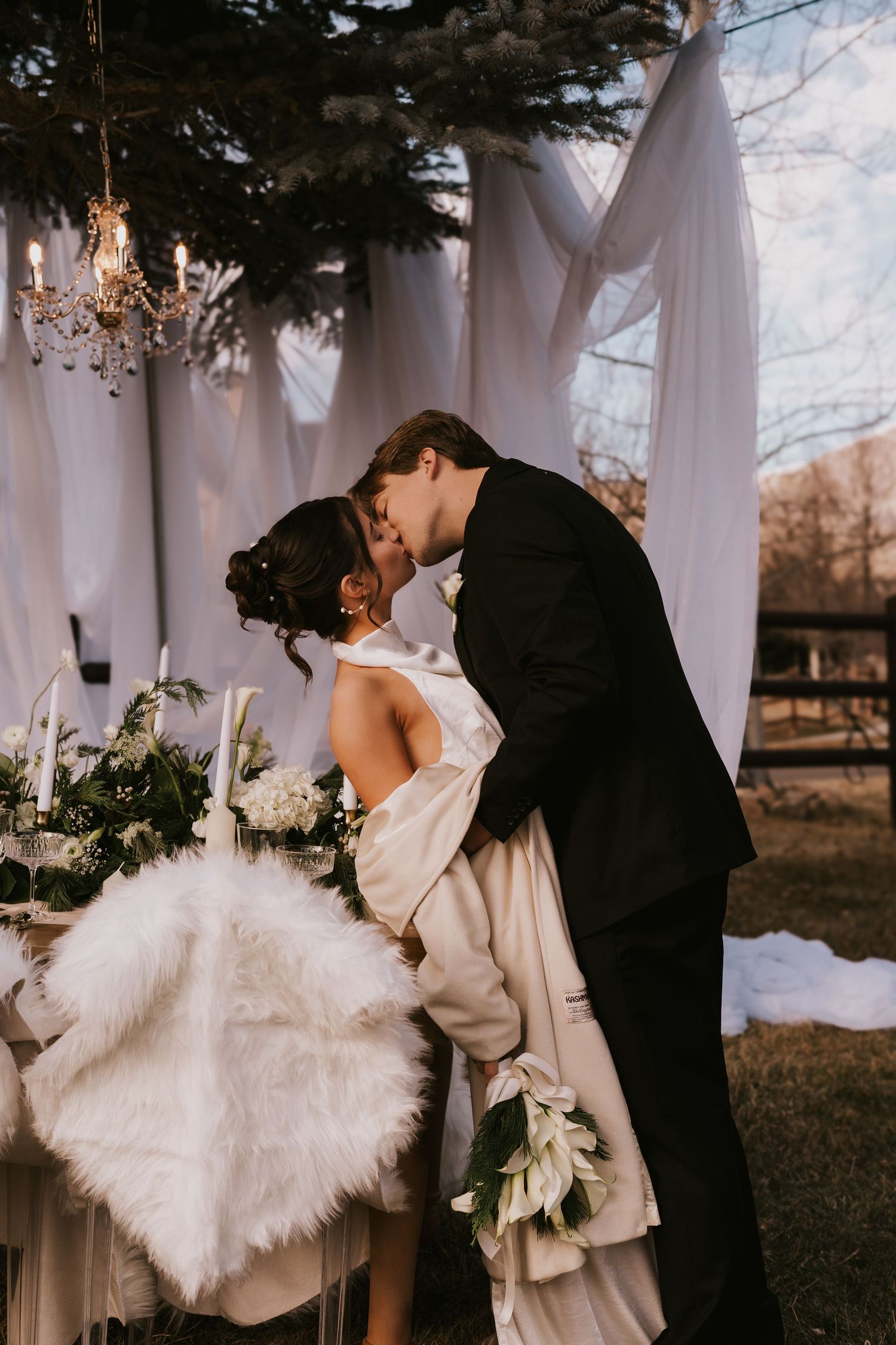 Romantic winter wedding photos showing couple embracing outdoors with white draping and evergreen trees in background.