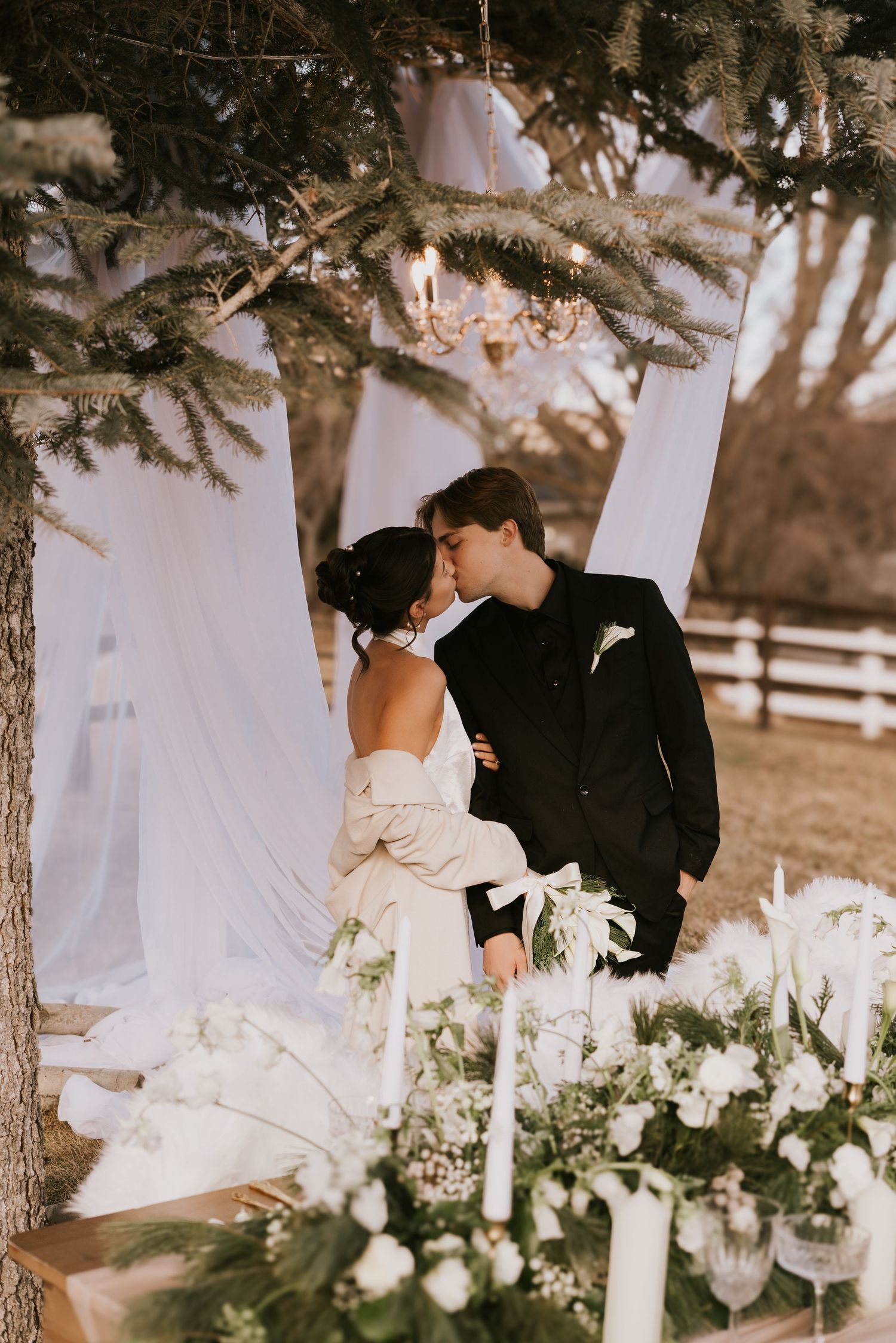 A romantic outdoor wedding scene with white floral arrangements and candles beneath evergreen trees at sunset.