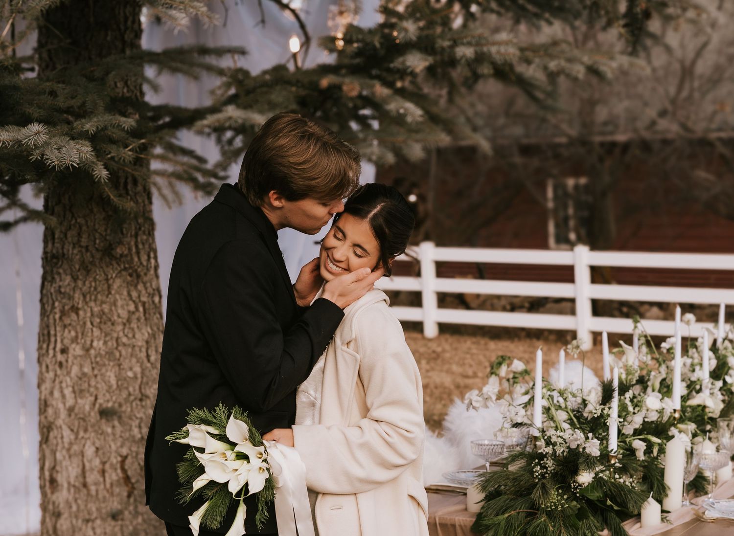 A romantic wedding couple embraces outdoors near a white gazebo decorated with floral arrangements.