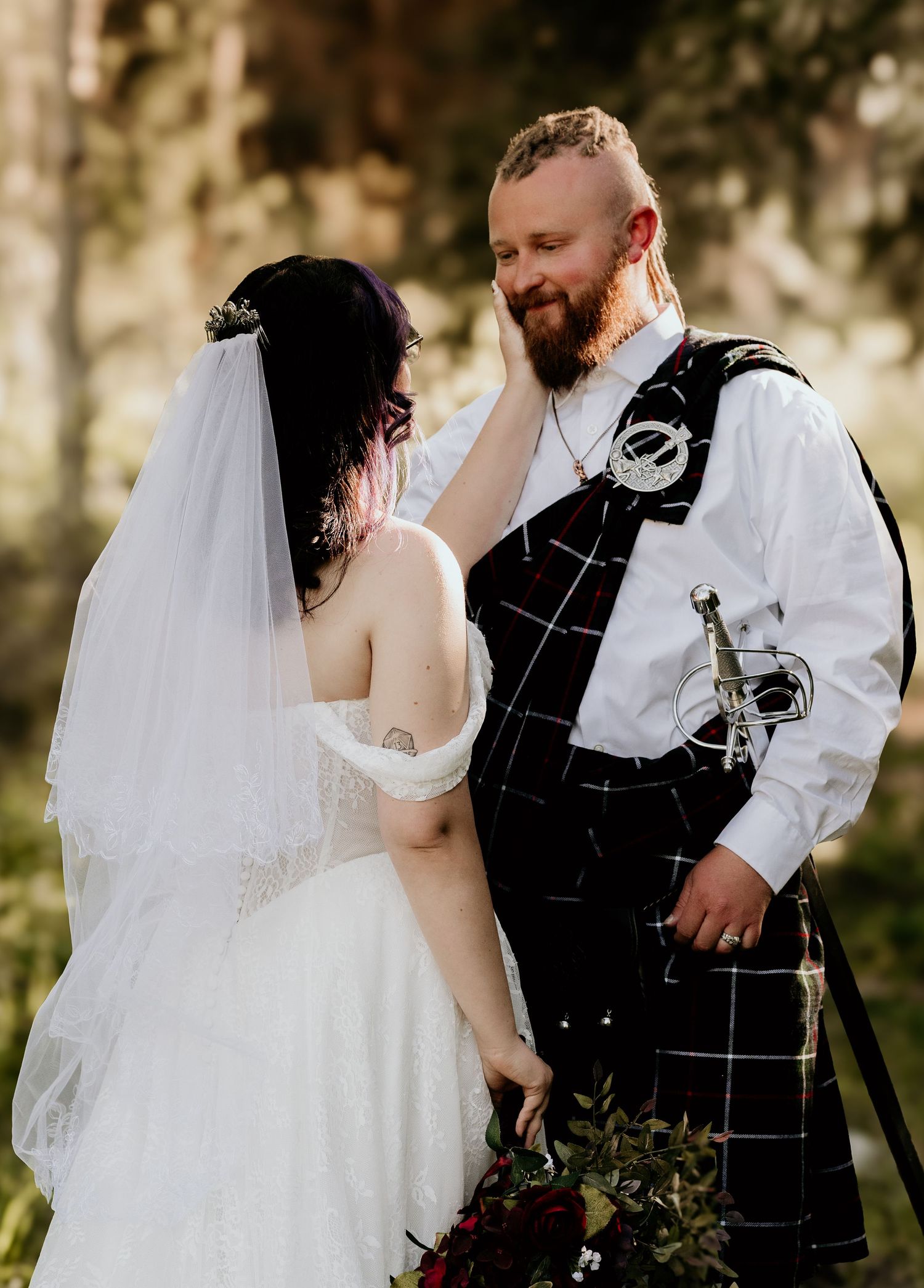 Bride in white gown and veil touches groom's face as he smiles, wearing a tartan kilt and sword outdoors.