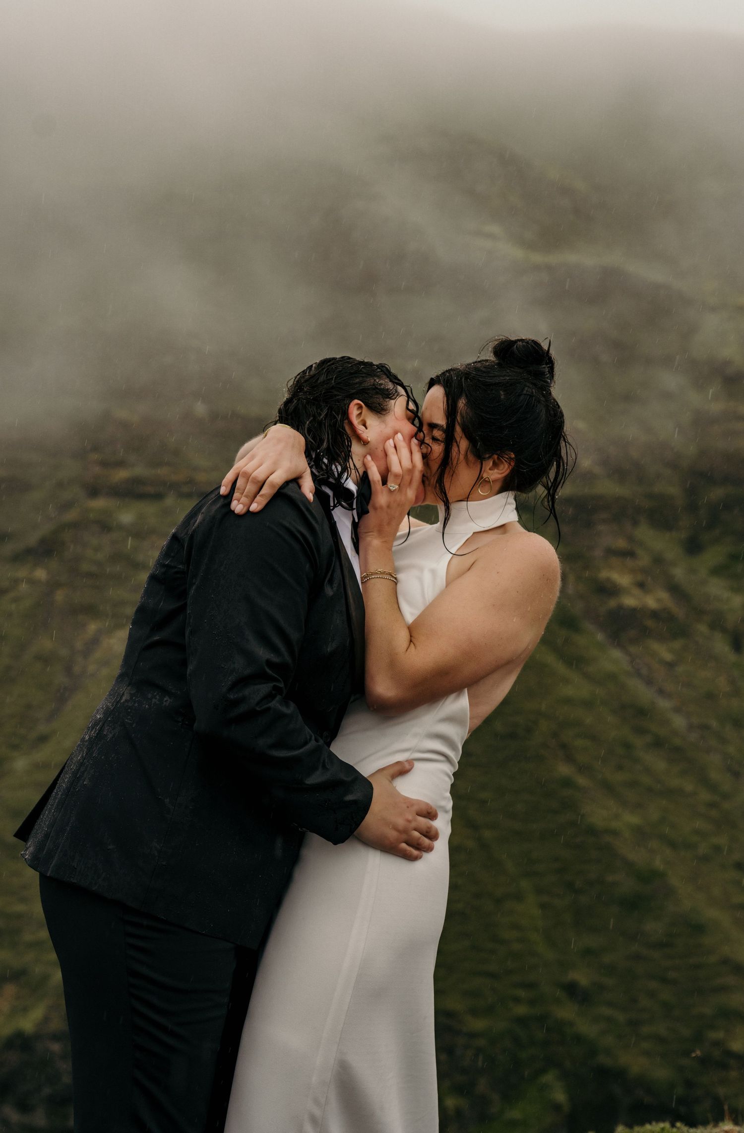 Two brides share a passionate kiss on a misty mountain overlook, one in a black suit and one in a white gown.