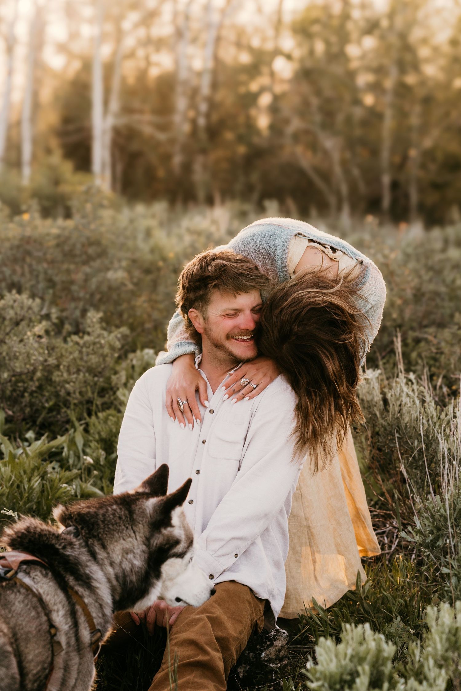 Couple laughing together in a meadow with a husky dog, surrounded by lush greenery and golden trees.