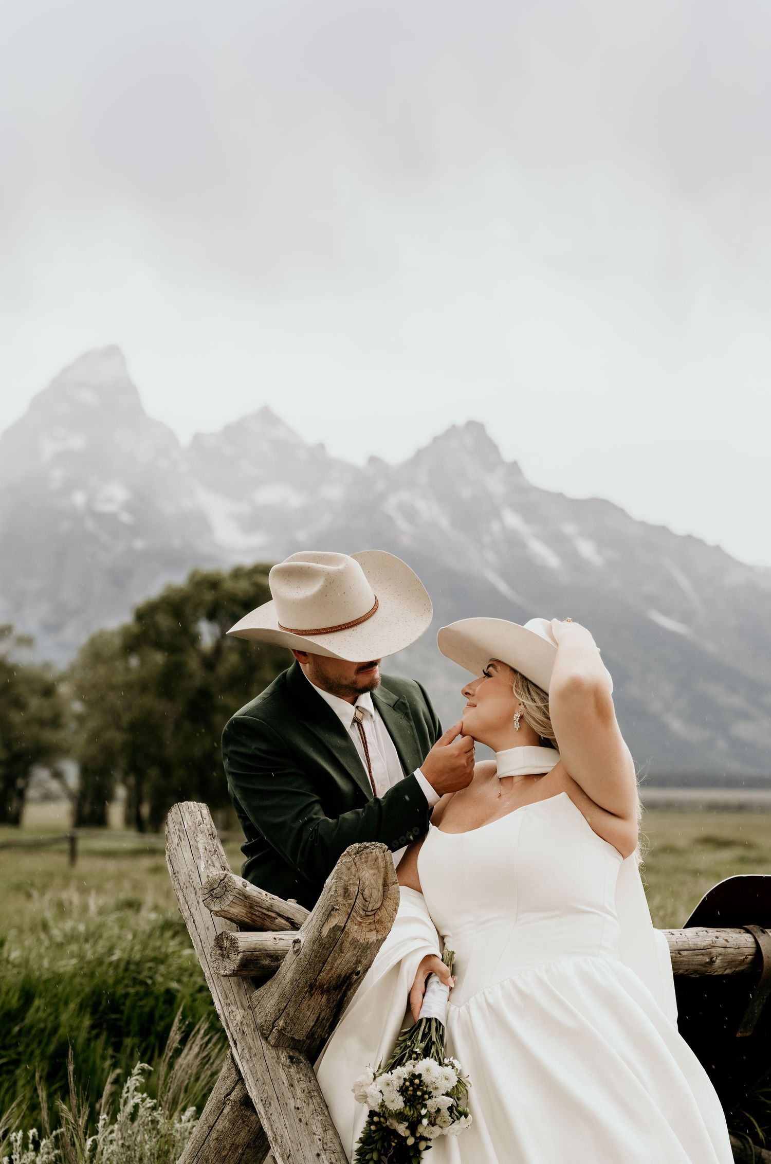 Couple in cowboy hats share a romantic moment by a rustic fence with Grand Teton mountains behind them.