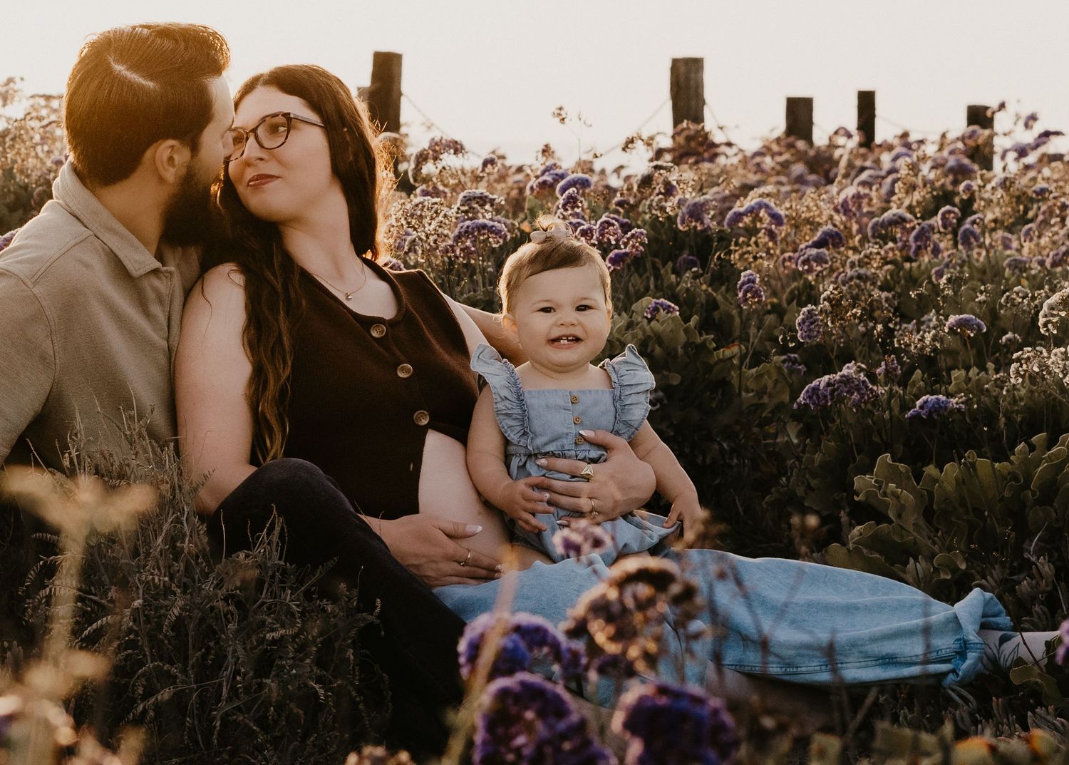 A young family sits together in a purple wildflower field at golden hour, with parents cradling their smiling baby.
