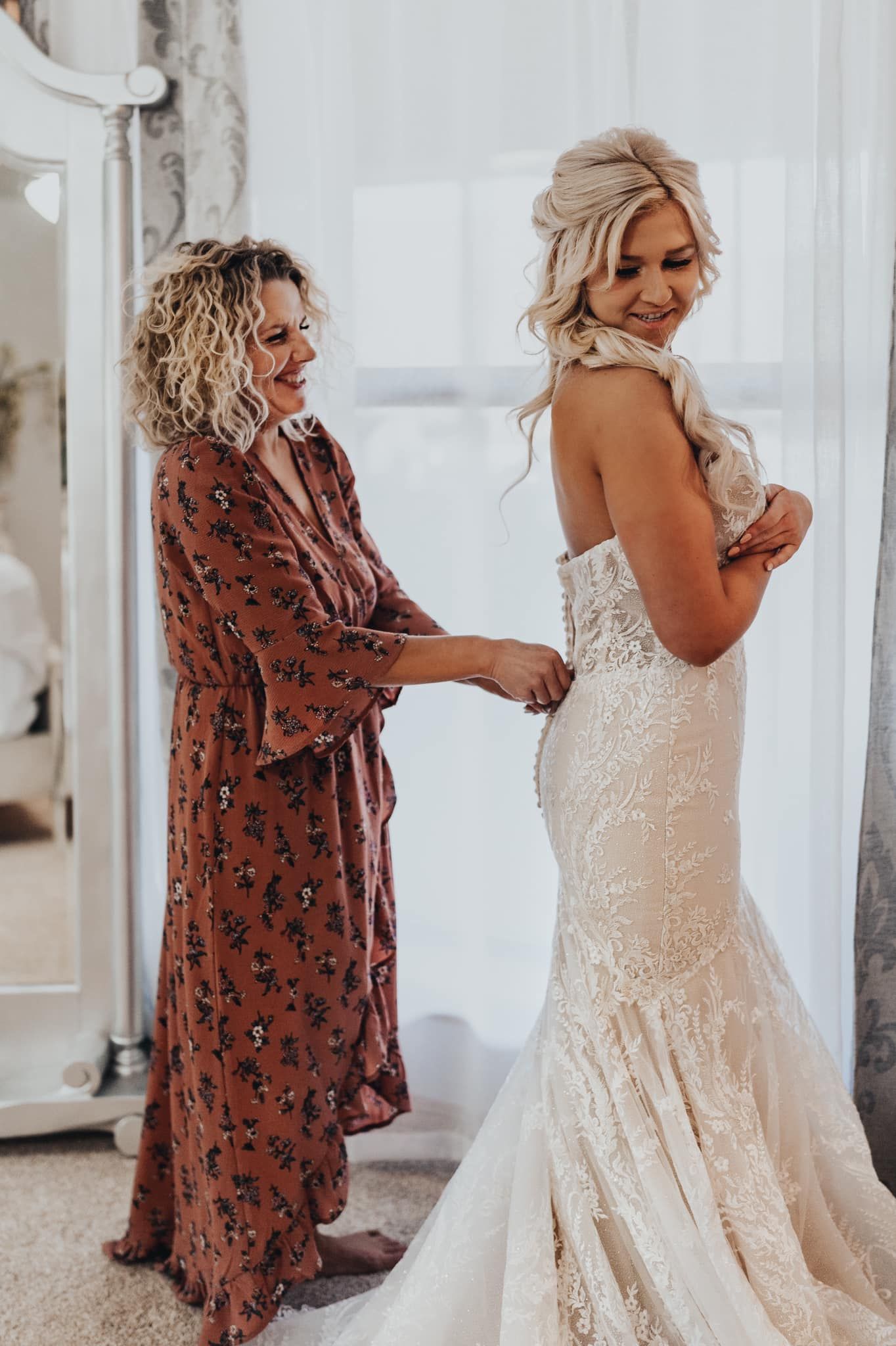 A woman in a floral dress helps adjust a stunning lace mermaid wedding gown in a bright sunlit room.